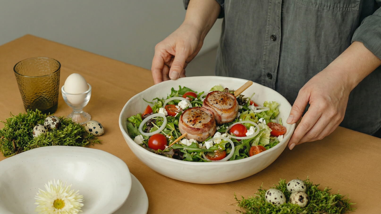 Eine Person präsentiert eine große Schüssel mit gemischtem Salat. Der Salat enthält grüne Blätter, Kirschtomaten, Zwiebelringe und Feta. Auf dem Salat liegen zwei mit Speck umwickelte Fleischscheiben. Im Hintergrund sind ein Glas, ein Ei und einige Wachteleier zu sehen.