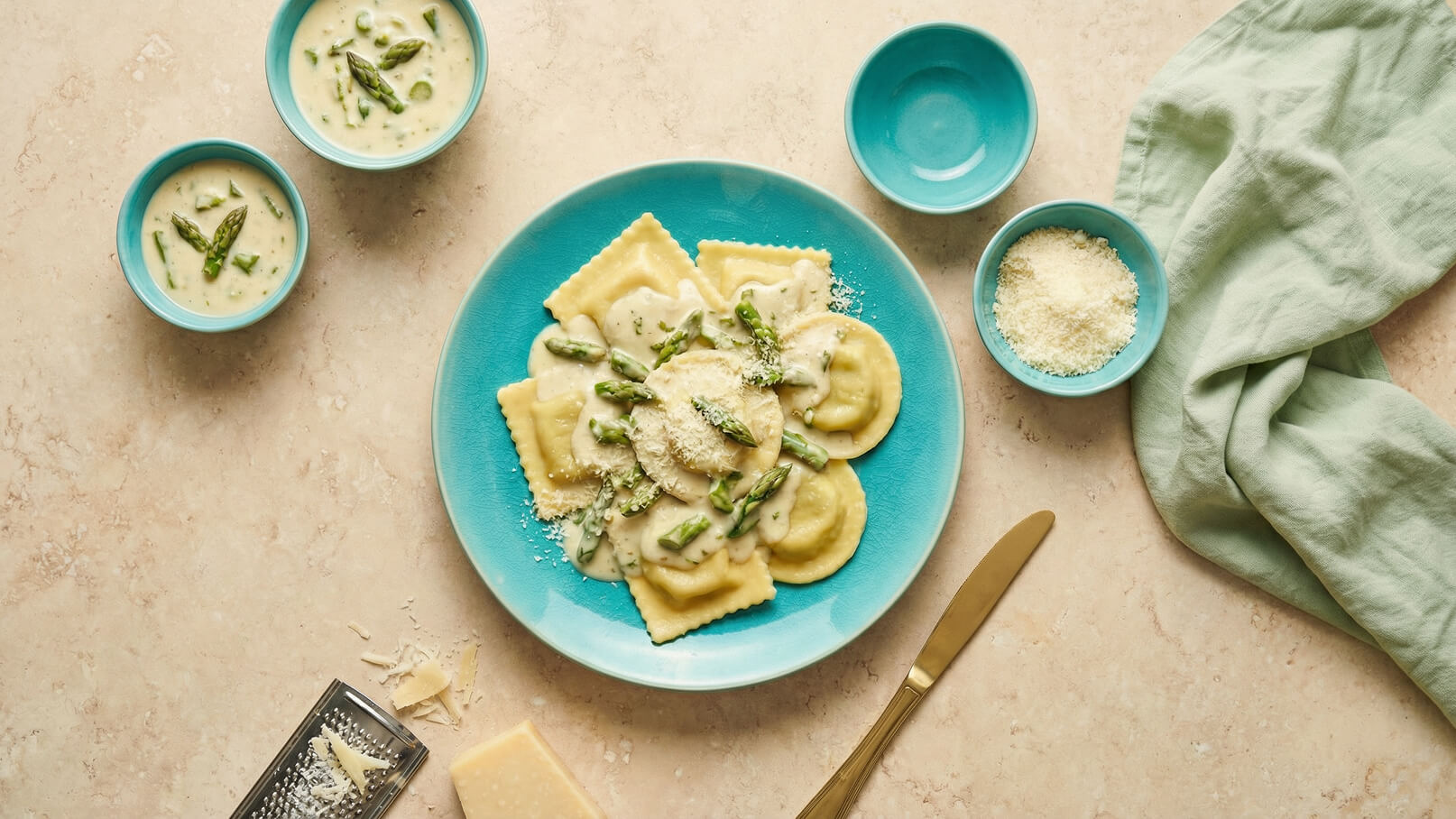 Ein blauer Teller mit Ravioli in einer cremigen Sauce, garniert mit grünen Spargelstücken und geriebenem Käse. Daneben befinden sich einige kleine Schalen mit einer weiteren Spargelsauce und geriebenem Käse, sowie ein Besteckset und ein Tuch.