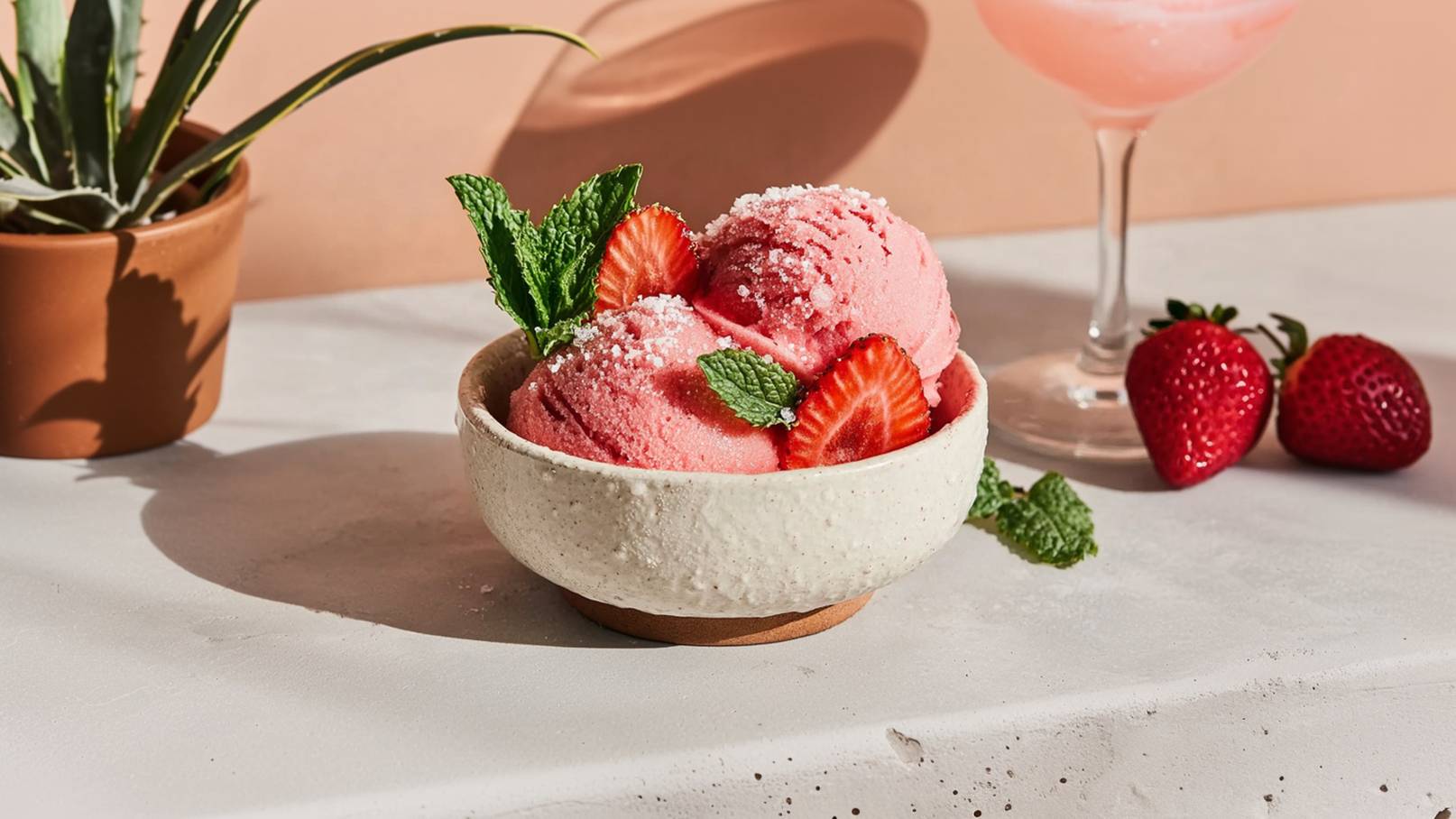 Schüssel mit erfrischendem Erdbeersorbet, garniert mit Minzblättern und Zitronenzesten. Um die Schüssel sind frische Erdbeeren und ein Zitronenschnitz angeordnet, auf einem marmorierten Tisch.