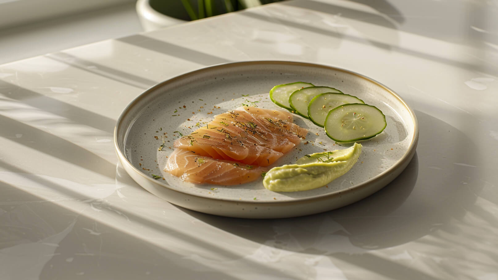 Ein eleganter Teller mit dünn geschnittenem Lachs, garniert mit Kräutern und Gewürzen. Daneben liegen einige Gurkenscheiben und ein Klecks grünes Püree. Der Teller steht auf einer hellen Oberfläche, die von sanften Schatten durchzogen ist.