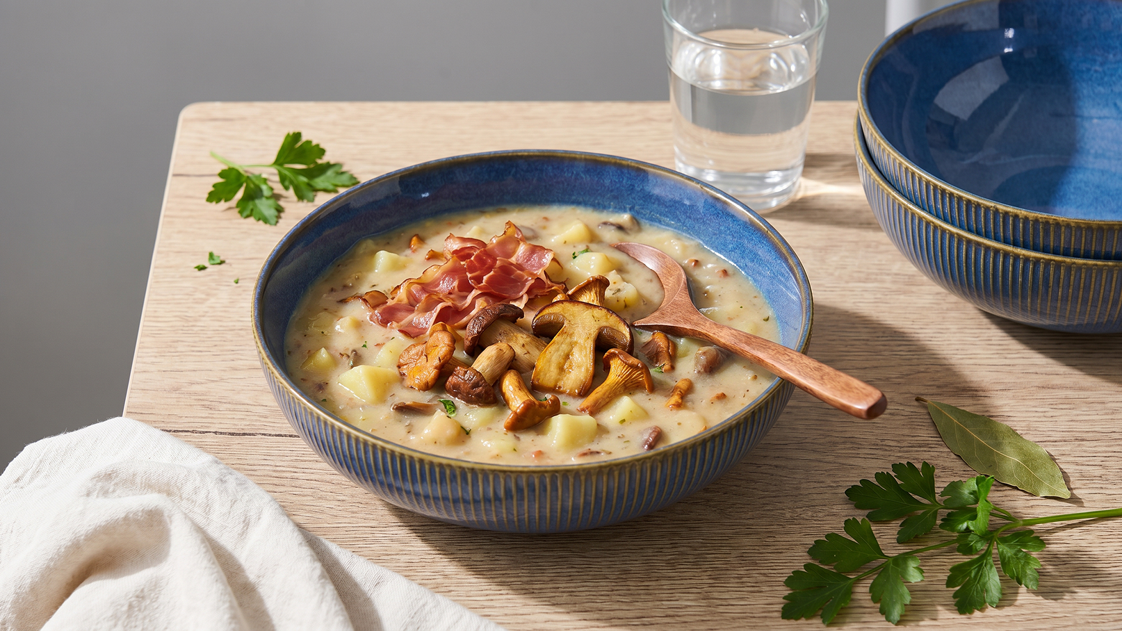 Eine Schüssel mit cremiger Pilzsuppe, garniert mit gebratenen Pilzen und Schinkenstreifen. Neben der Schüssel liegt ein Holzlöffel. Im Hintergrund steht ein Glas Wasser und mehrere leere Schalen. Frische Petersilie liegt dekorativ auf dem Tisch.