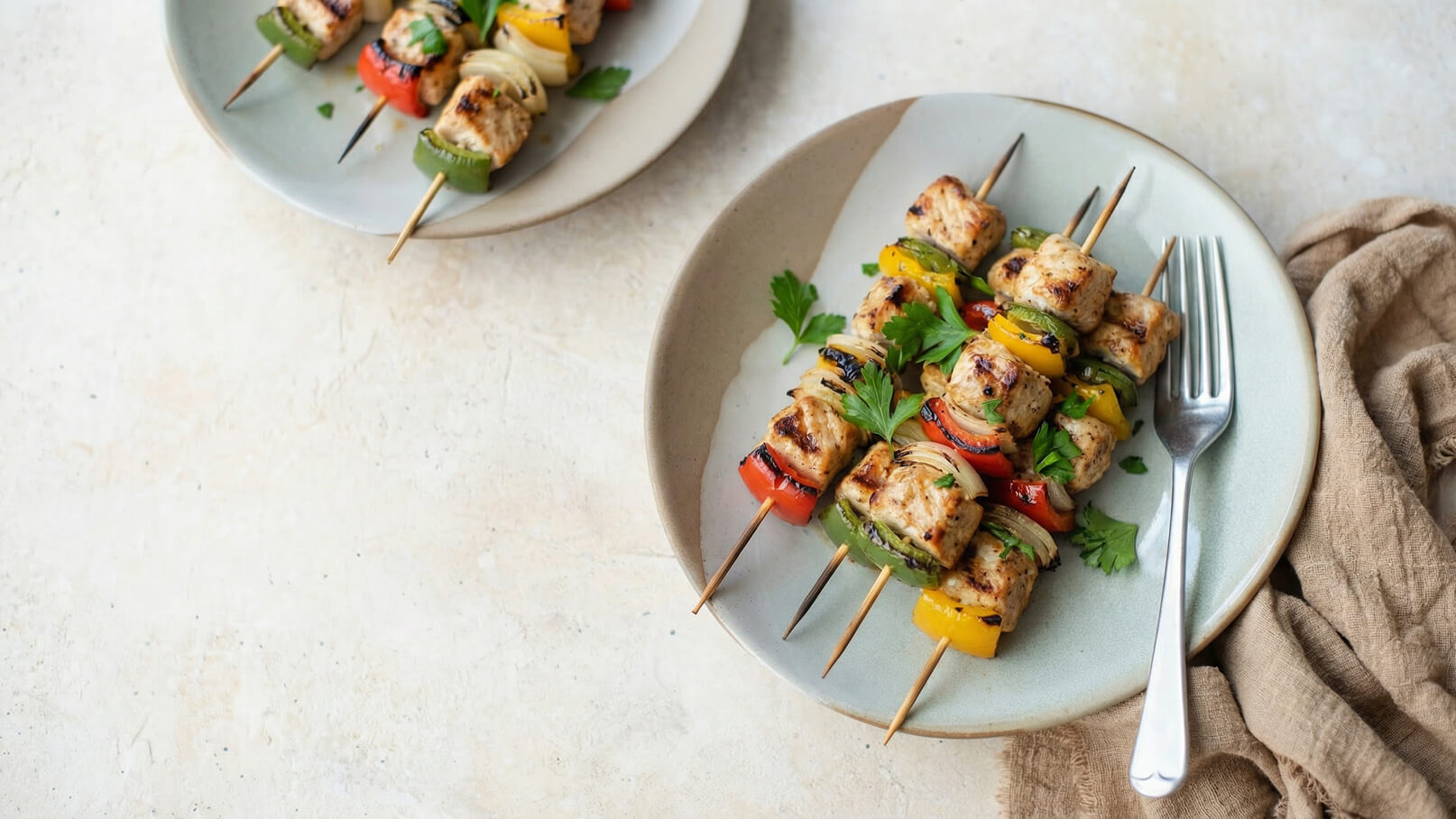 Auf dem Bild sind gegrillte Spieße mit Fleisch und buntem Gemüse zu sehen, serviert auf einem hellen Teller. Die Spieße sind mit grünen, roten, gelben Paprika und Zwiebeln garniert. Im Hintergrund steht ein zweite Teller mit weiteren Spießen und einer Gabel.
