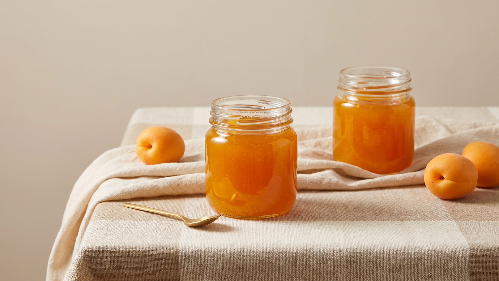 Zwei Gläser mit goldener Aprikosenmarmelade stehen auf einem Tisch mit einer gestreiften Tischdecke. Neben den Gläsern liegen frische Aprikosen und ein Löffel aus Metall. Die Szene vermittelt einen einladenden, frischen Eindruck.