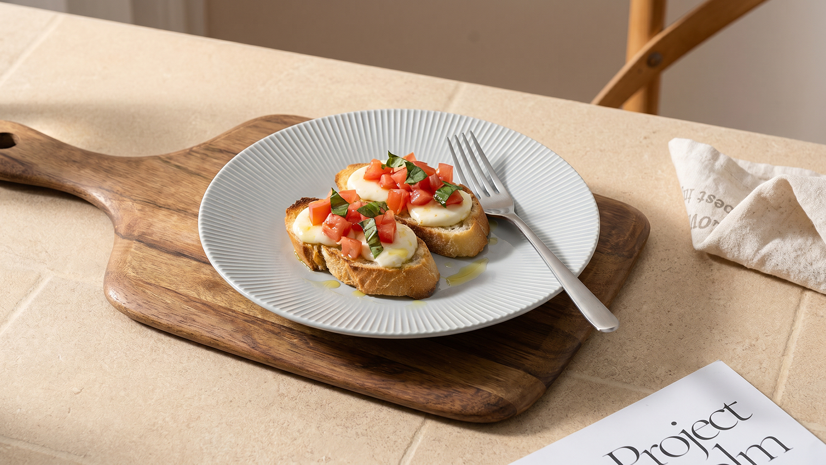 Ein Teller mit geröstetem Brot belegt mit frischen Tomaten und Basilikum befindet sich auf einem Holzbrett. Eine Gabel liegt neben dem Teller. Im Hintergrund ist ein Tisch und eine Serviette zu sehen.