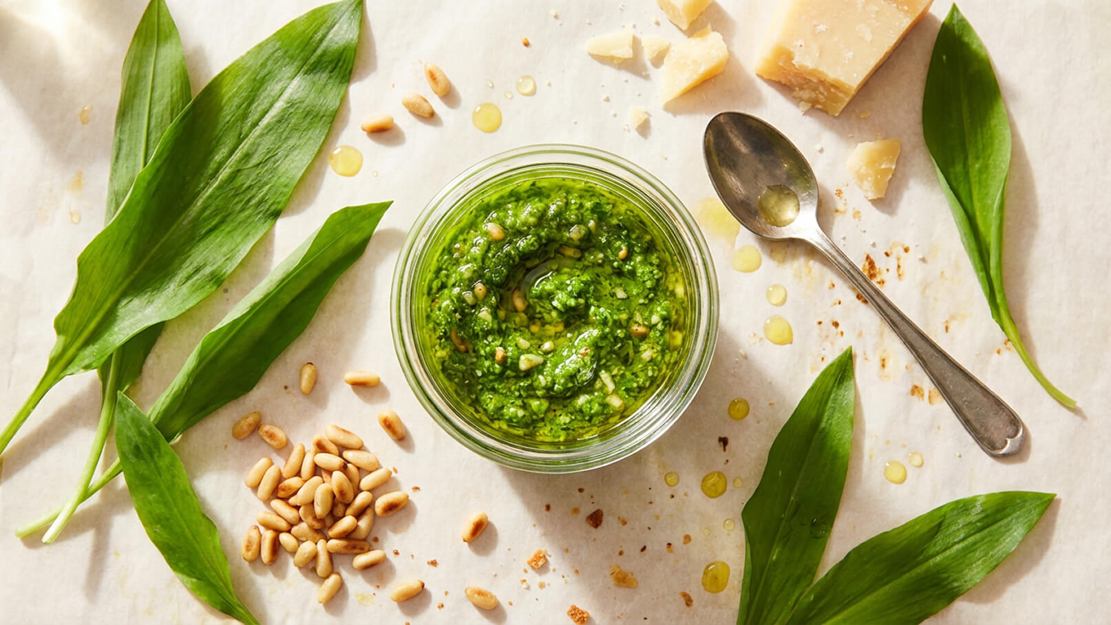 Ein Glas Bärlauchpesto liegt auf Backpapier, umgeben von frischen Bärlauchblättern, Pinienkernen und einigen Käsestücken. Im Hintergrund sind Tropfen von Olivenöl sichtbar. Der Fokus liegt auf der leuchtend grünen Pesto-Masse im Glas.