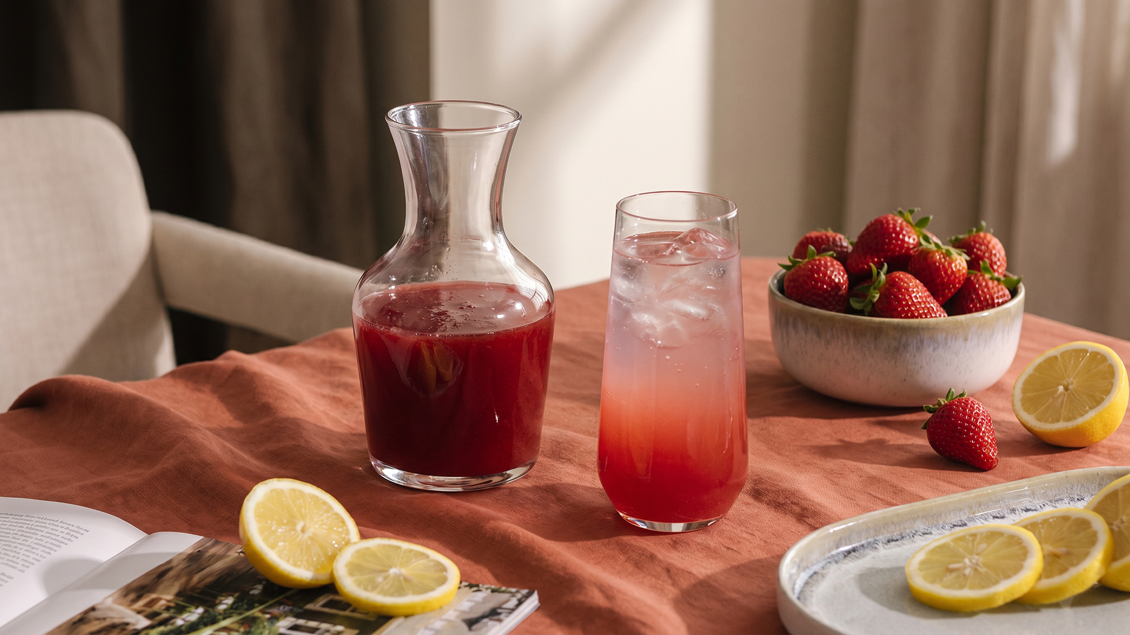 Auf einem Tisch mit orangefarbenem Tuch stehen eine Glaskaraffe mit rotem Getränk und ein Glastumbler mit eisgekühltem, rotem Getränk. Neben ihnen liegt ein Buch, und eine Schüssel mit frischen Erdbeeren sowie einige Zitronenscheiben sind ebenfalls sichtbar.