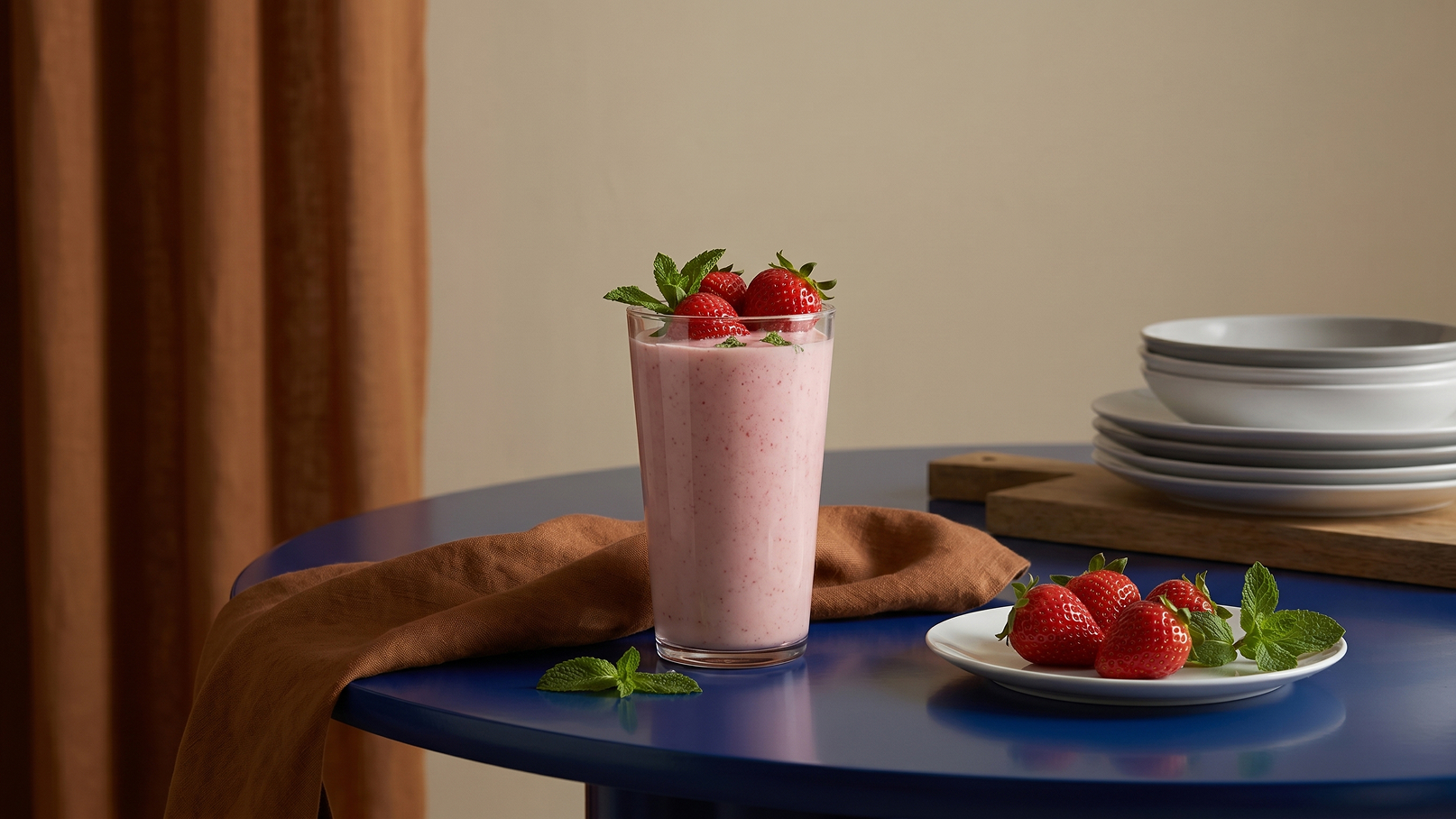 Ein großes Glas Erdbeersmoothie steht auf einem blauen Tisch, garniert mit frischen Erdbeeren und einem Minzblatt. Daneben liegt ein Teller mit weiteren Erdbeeren. Im Hintergrund sind stapelbare Geschirrteile sichtbar. Die Atmosphäre ist freundlich und einladend.