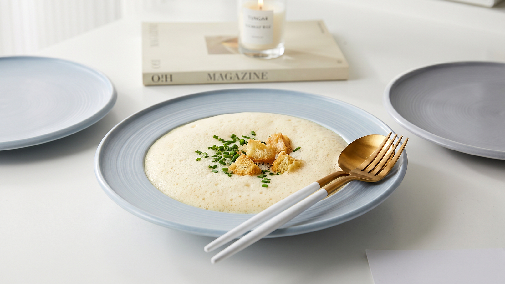 Eine helle, cremige Suppe ist in einem runden, blauen Teller angerichtet und mit gehacktem Schnittlauch und knusprigen Croutons garniert. Rechts liegt Besteck mit einer goldenen Gabel und einer weißen Esslöffel. Im Hintergrund sind zwei graue Teller und eine Kerze auf einem Tisch sichtbar.
