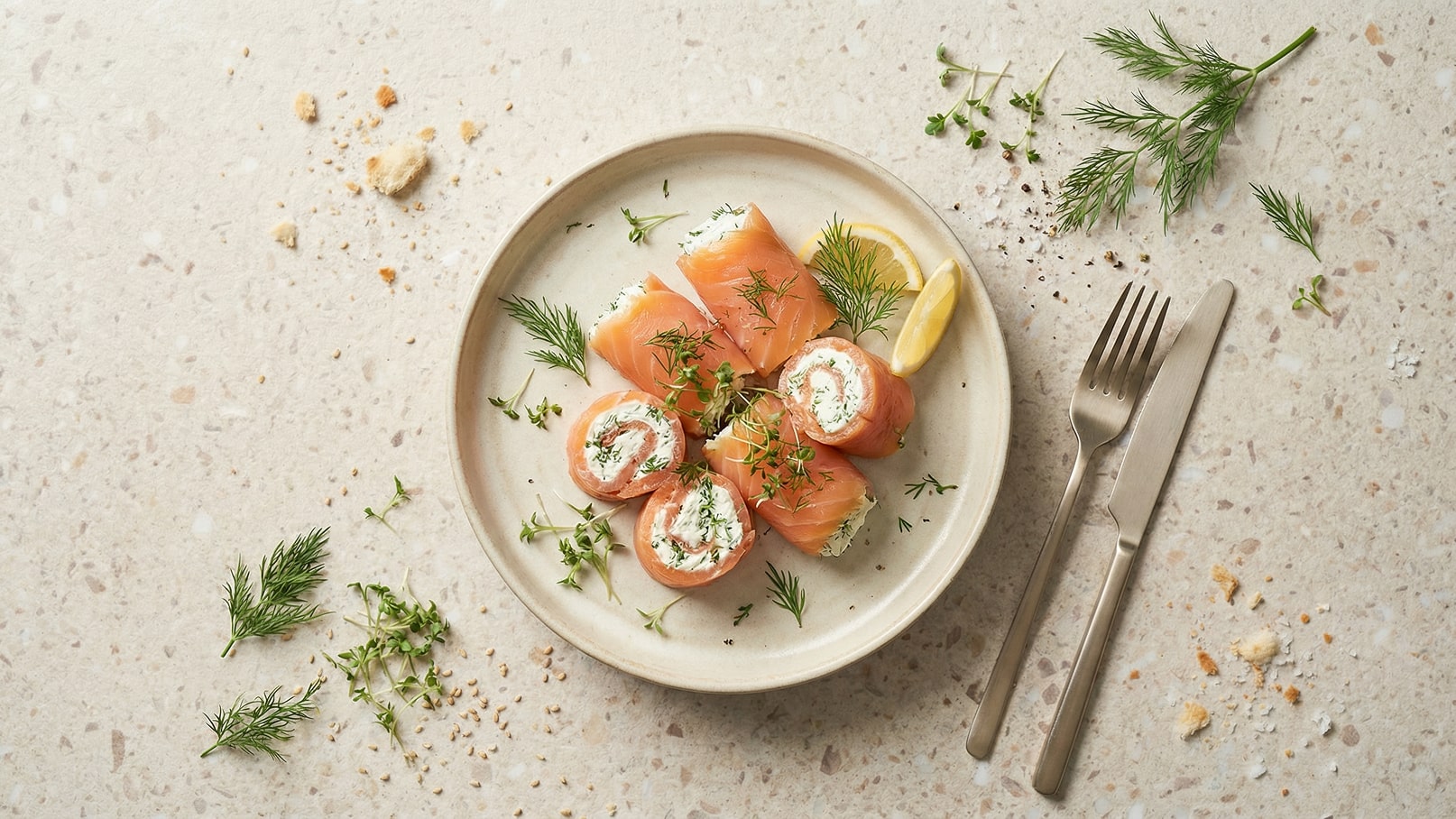 Ein Teller mit verschiedenen Sushi-Rollen, gefüllt mit Frischkäse und umhüllt von Lachs. Dazu frische Kräuter wie Dill und Zitronenscheiben. Besteck liegt daneben.