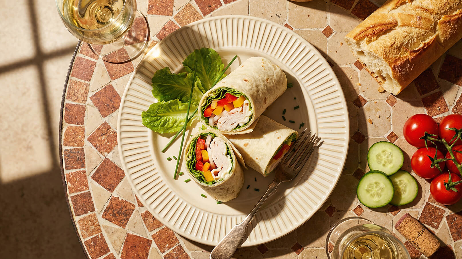 Auf einem Teller liegen zwei aufgerollte Tortillas mit gefülltem Gemüse und Putenfleisch. Daneben befindet sich ein Blatt grüner Salat und Schnittlauch. Im Hintergrund sind einige Kirschtomaten und Gurkenscheiben zu sehen. Ein Glas Wein steht ebenfalls auf dem Tisch.