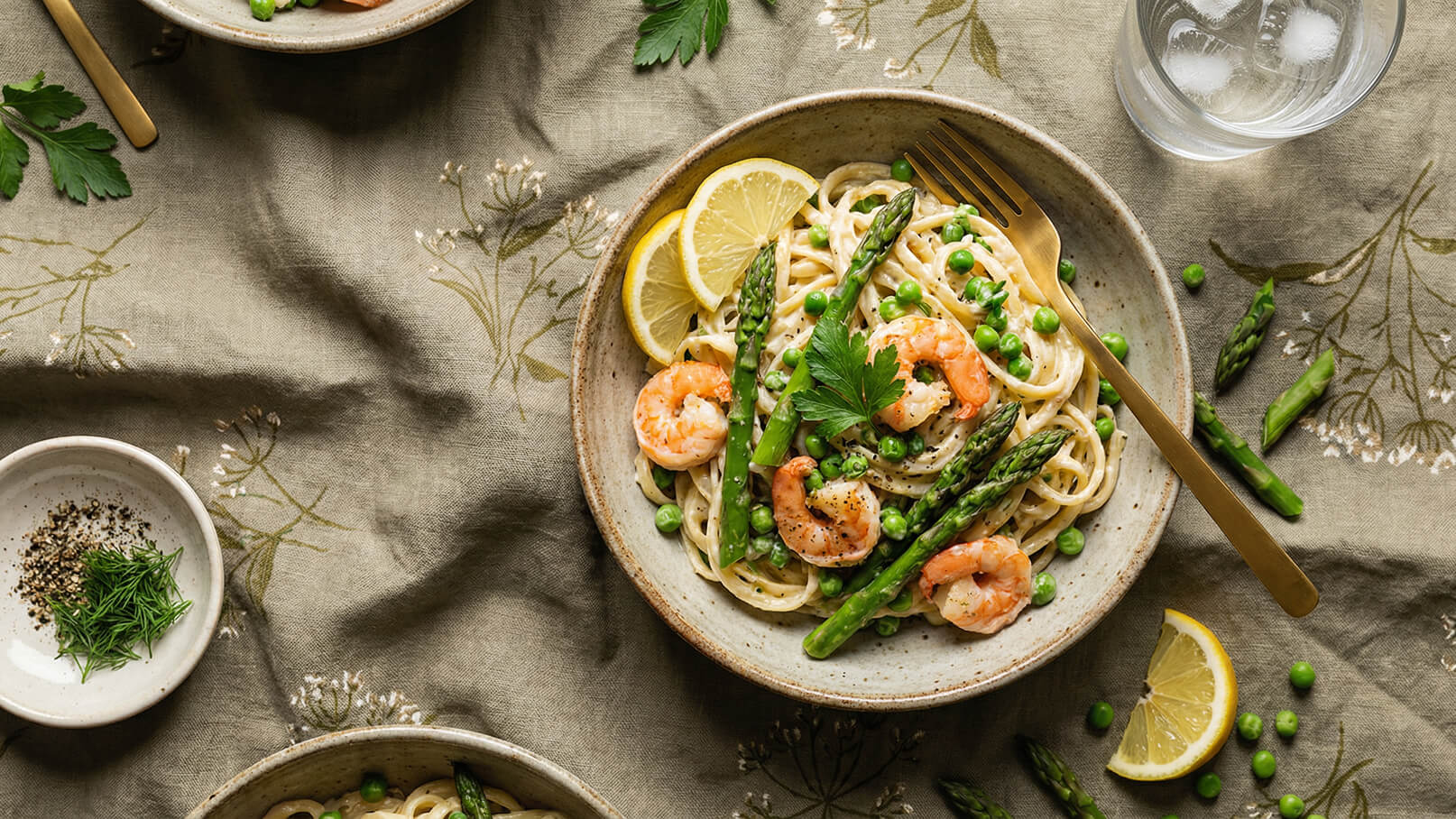 Ein Teller Pasta mit Garnelen, grünem Spargel und Erbsen. Die Speise ist mit frischen Kräutern und Zitronenscheiben garniert. Im Hintergrund sind eine Gabel und ein Glas Wasser zu sehen. Der Tisch ist mit einer dezenten, gemusterten Tischdecke bedeckt.