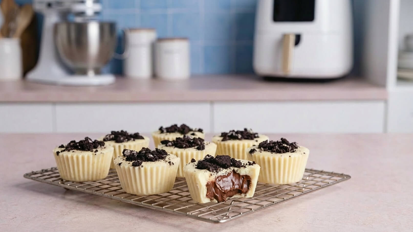Neun weiße Schokoladen-Muffins auf einem Kuchengitter, gefüllt mit einer schokoladigen Creme und bedeckt mit zerbröselten Oreos. Einer der Muffins ist angeschnitten, sodass die Füllung sichtbar ist. Im Hintergrund stehen Küchengeräte und Aufbewahrungsgläser.
