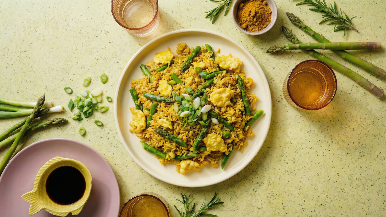 Ein Teller mit gebratenem Reis, der mit grünem Spargel und Rührei garniert ist. Frisch geschnittene Frühlingszwiebeln liegen darüber. Um den Teller herum befinden sich Gewürze und Getränke, die zur Essenspräsentation beitragen. Der Hintergrund ist in sanften Grüntönen gehalten.