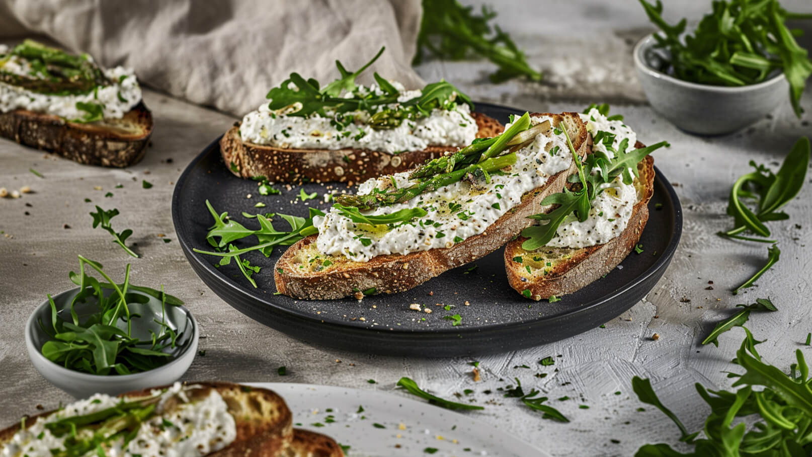 Ein schwarzes Teller mit mehreren Scheiben knusprigem Brot, belegt mit einer cremigen Käseaufstrich und frischen grünen Spargelstücken. Darüber sind gehackte Kräuter und Rucola verteilt. Im Hintergrund ist eine kleine Schüssel mit frischem Rucola zu sehen.