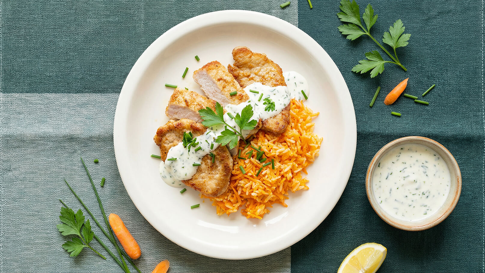 Ein Teller mit gebratenem Hähnchen, garniert mit einer weißen Sauce und frischer Petersilie, liegt auf einem Bett aus orangefarbenem Reis. Daneben gibt es eine Schale mit zusätzlicher Sauce, einen Zitronenschnitz und einige frische Kräuter sowie Karottenstücke.