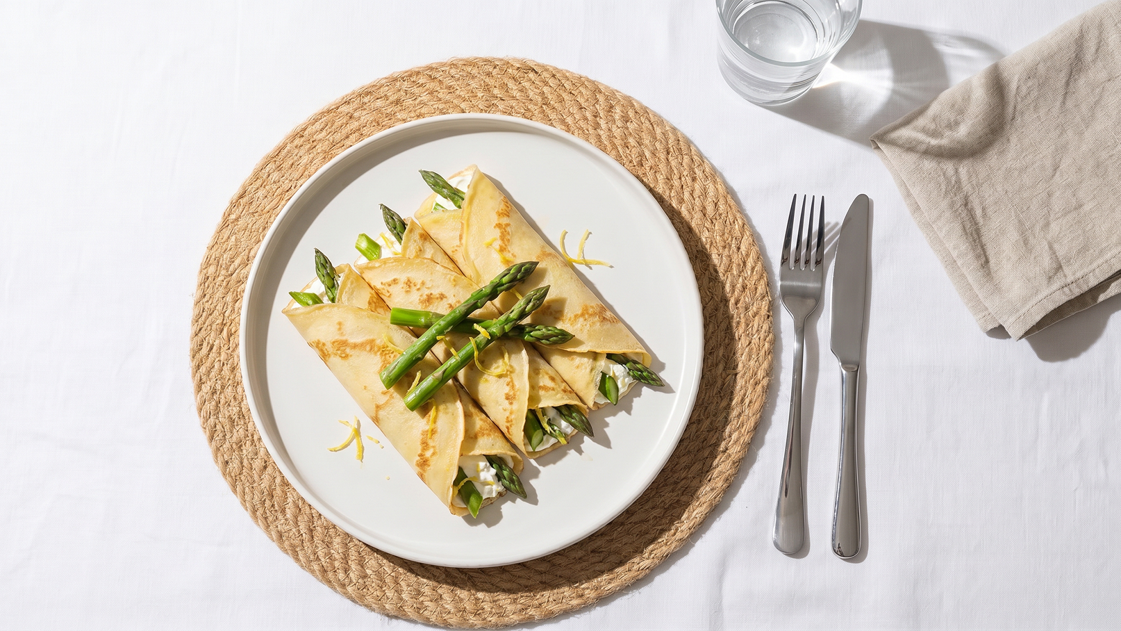 Ein weißer Teller auf einem runden Untersetzer, gefüllt mit gefüllten Crêpes. Die Crêpes sind mit Spargel und frischem Grün garniert. Daneben sind Besteck und ein Glas Wasser sichtbar.