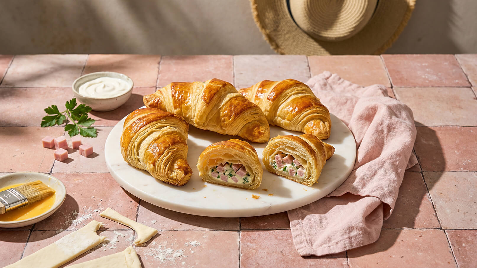 Ein runder Teller mit mehreren goldbraunen Croissants, von denen eines aufgeschnitten ist und gefüllt mit Schinken und Kräutern sichtbar ist. Der Hintergrund zeigt eine rustikale Tischoberfläche und einige Zubereitungselemente.
