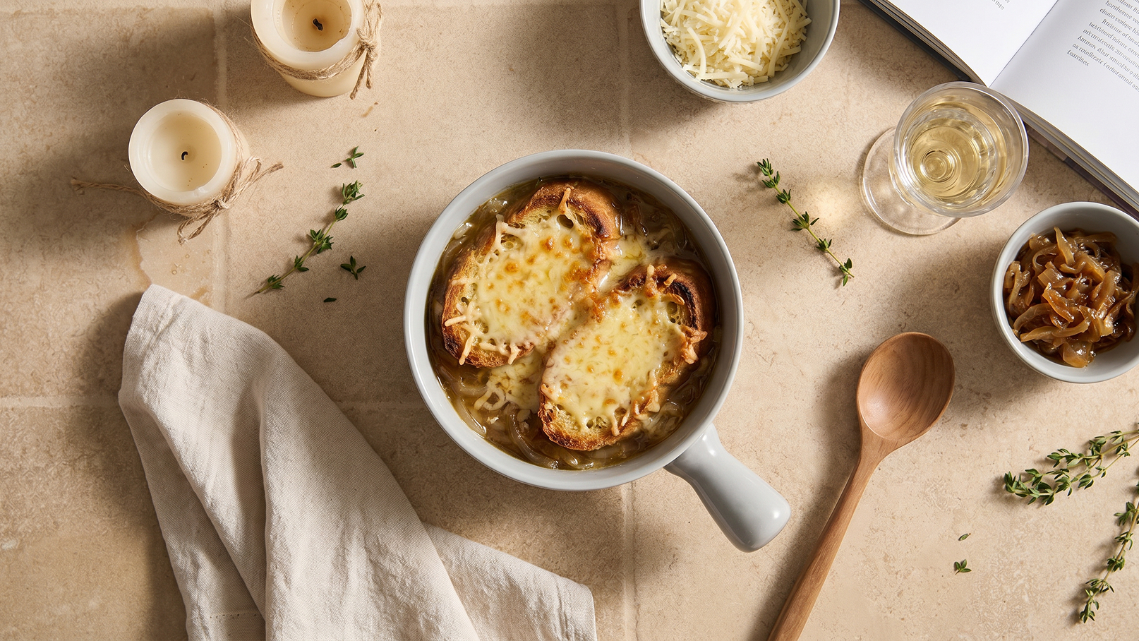 Eine Schale mit französischer Zwiebelsuppe, belegt mit geschmolzenem Käse und zwei gerösteten Brotscheiben. Umgeben von Kräutern, einer Serviette, Kerzen und einem Glas Weißwein. Ein Kochbuch steht offen im Hintergrund.