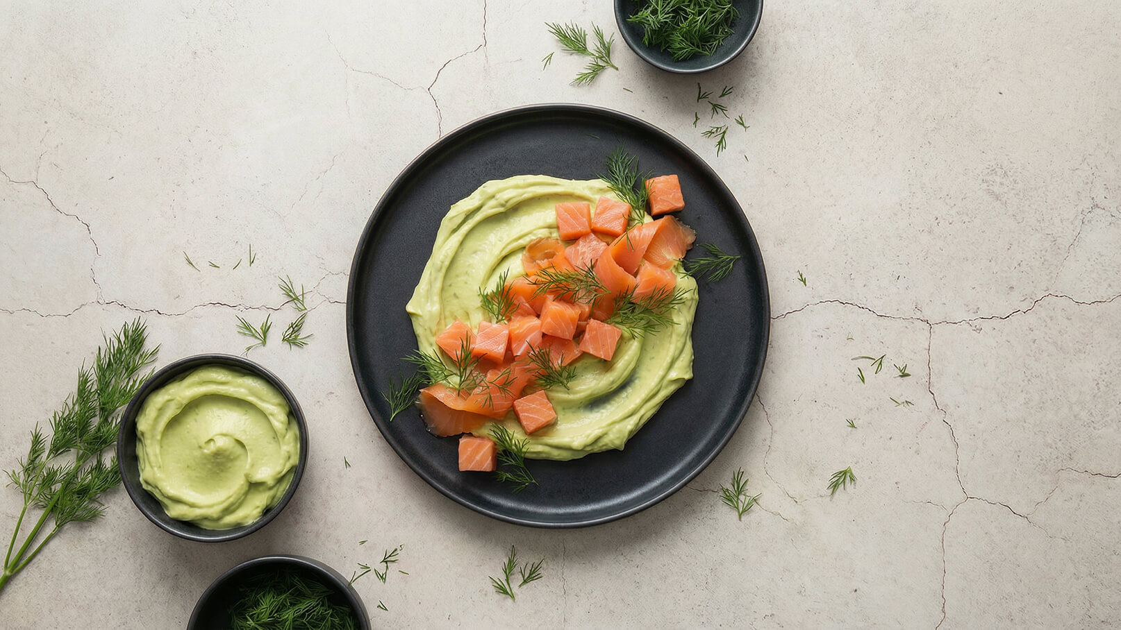 Ein schwarzer Teller mit cremigem Avocado-Dip, garniert mit würfelig geschnittenem Lachs und frischem Dill. Daneben befinden sich zwei Schalen mit ähnlichem Dip und etwas Dill. Der Untergrund ist hell und hat feine Risse.