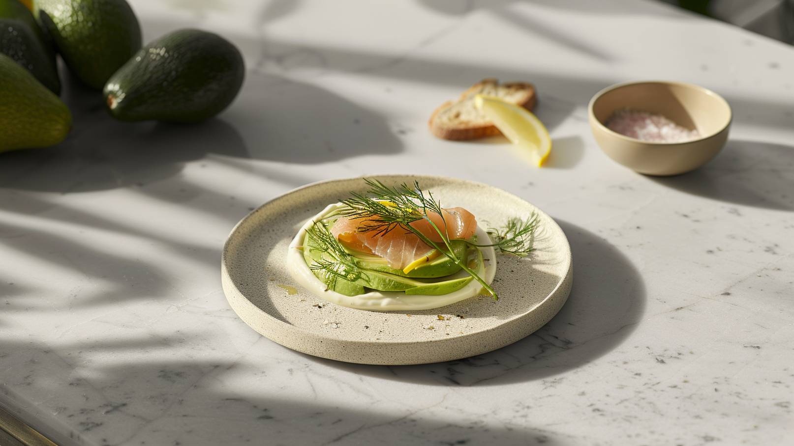 Ein Teller mit frischem, zartem Lachs, umgeben von grünen Bohnen und einer Schicht mildem Joghurt. Ein Zweig Dill ziert die oberste Schicht. Im Hintergrund sind Avocados und eine Schale mit Salz sichtbar.