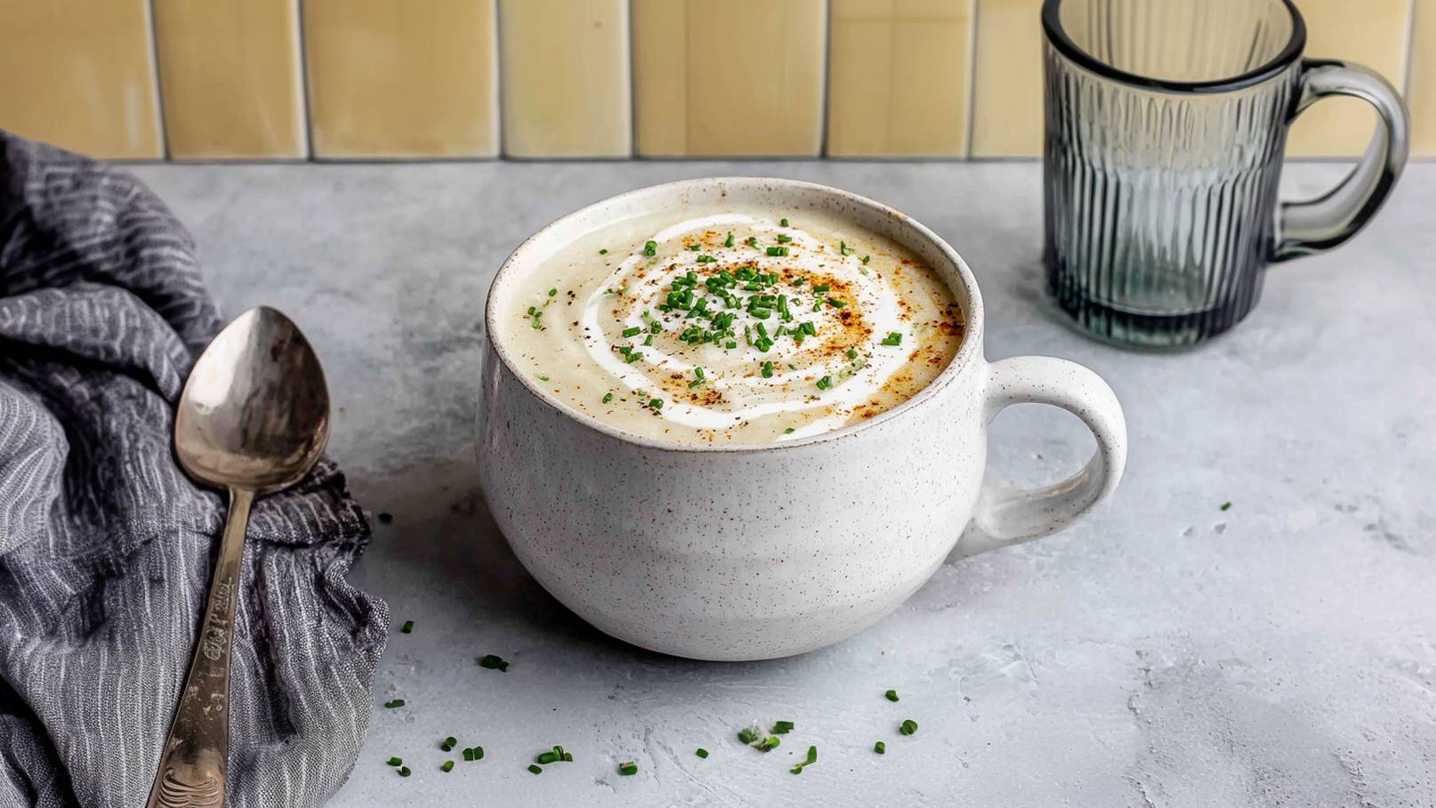 Eine große, weiße Schüssel mit cremiger Suppe, dekoriert mit einem Sahnehäufchen und Schnittlauch. Um die Schüssel liegen grüne Schnittlauchröllchen und etwas Paprika. Im Hintergrund steht ein Glas mit einem schlichtem Design.