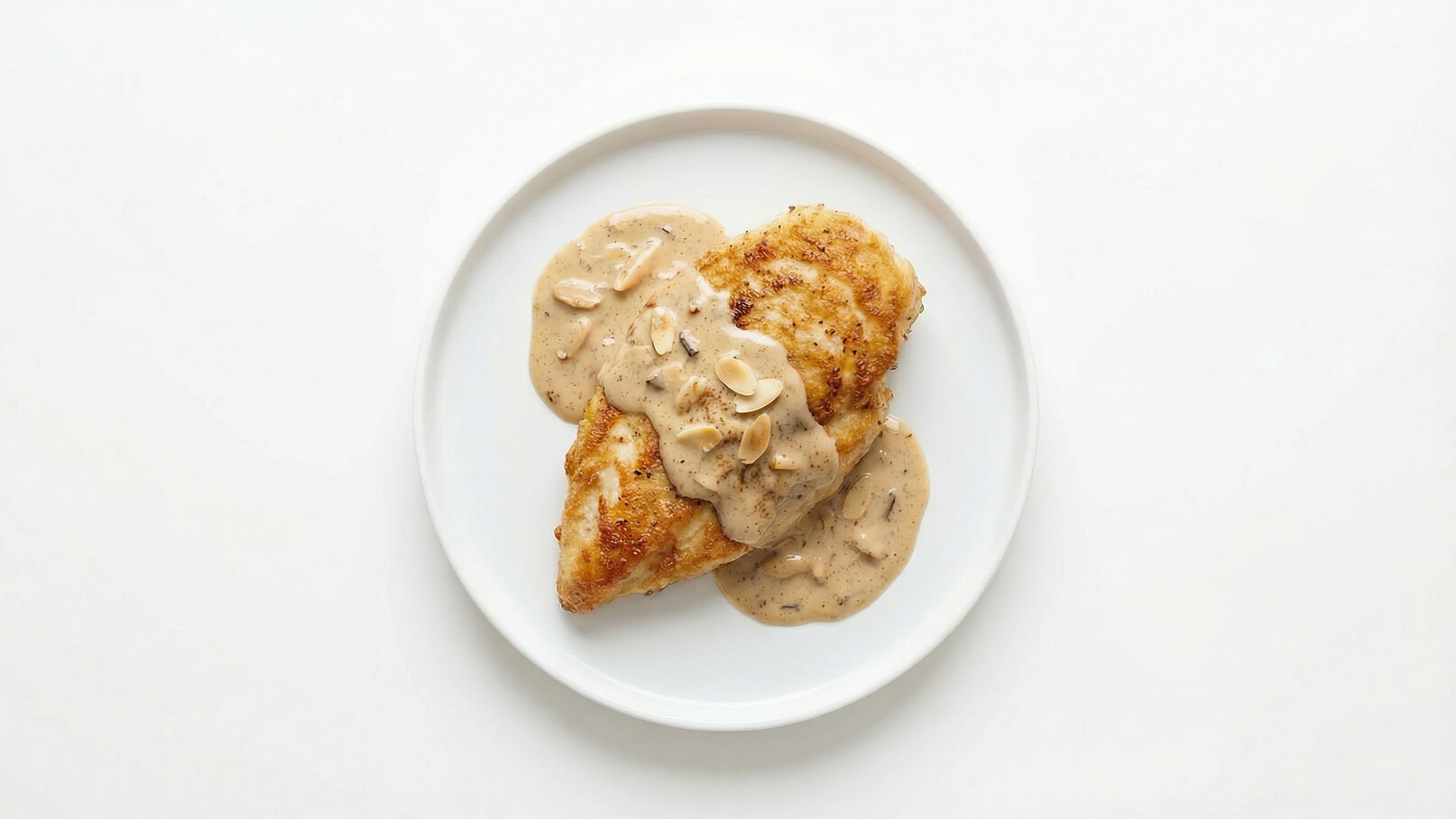 Ein Teller mit zwei knusprigen Hähnchenfilets, die mit einer cremigen Sauce überzogen sind. Die Sauce ist hell und enthält kleine Stücke, möglicherweise Mandeln. Der Hintergrund ist weiß und schlicht, um das Gericht hervorzuheben.