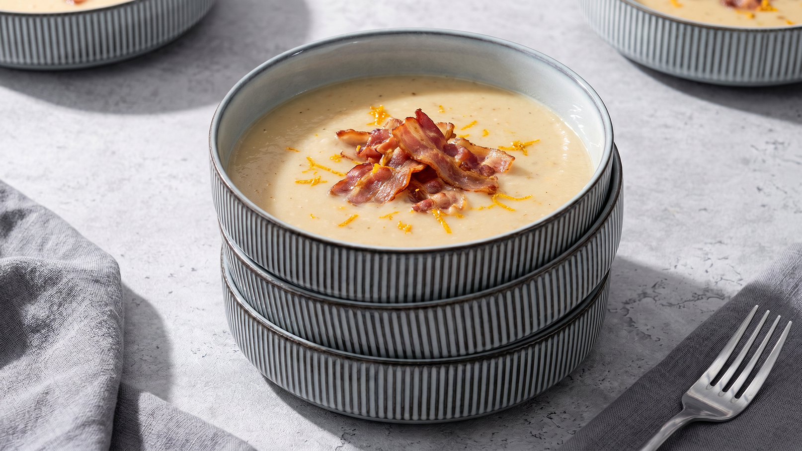 Eine gestapelte Anordnung von Schalen mit cremiger Suppe, die mit knusprigem Bacon und einer Zitronenabrieb-Dekoration serviert wird. Der Hintergrund zeigt eine neutrale Oberfläche mit einem Löffel und einem grauen Tuch.