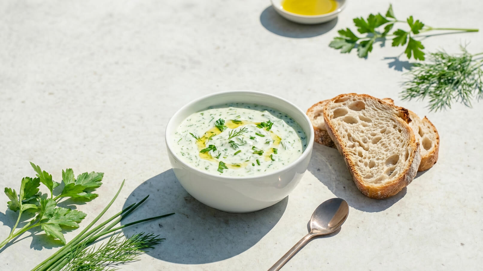 Eine Schüssel mit cremiger, grüner Kräutersuppe steht auf einem hellen Tisch. Neben der Schüssel liegen mehrere Scheiben Brot und einige frische Kräuter wie Petersilie und Dill. Ein Teelöffel ist ebenfalls zu sehen.