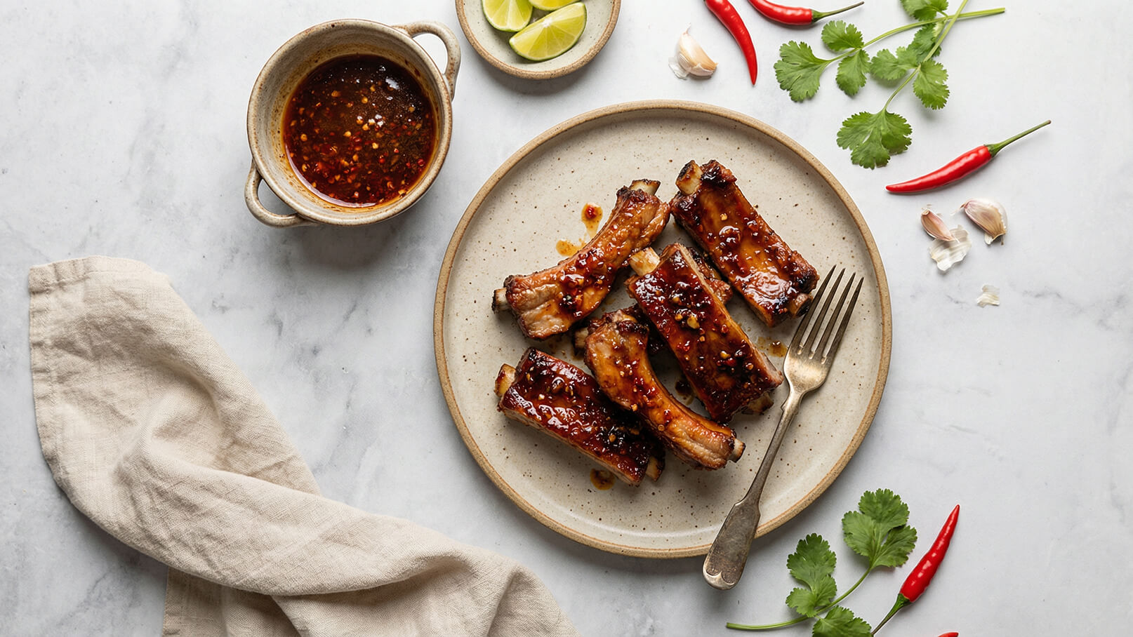 Ein Teller mit marinierten Rippchen ist in der Mitte abgebildet, daneben eine Schüssel mit einer würzigen Sauce. Frische Limettenscheiben, rote Chilis, Knoblauchzehen und Korianderblätter ergänzen das Gericht. Ein Besteckstück liegt auf dem Teller. Der Hintergrund ist marmoriert und hell.