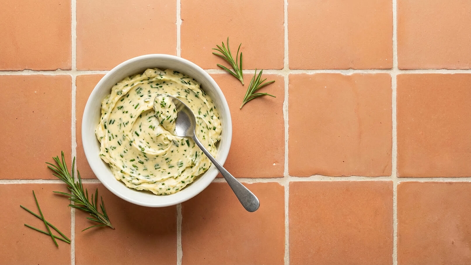 Eine weiße Schüssel enthält cremige Kräuterbutter, die mit einem Löffel leicht verrührt wurde. Daneben liegen einige frische Rosmarinnadeln auf einem terrakottafarbenen Untergrund.