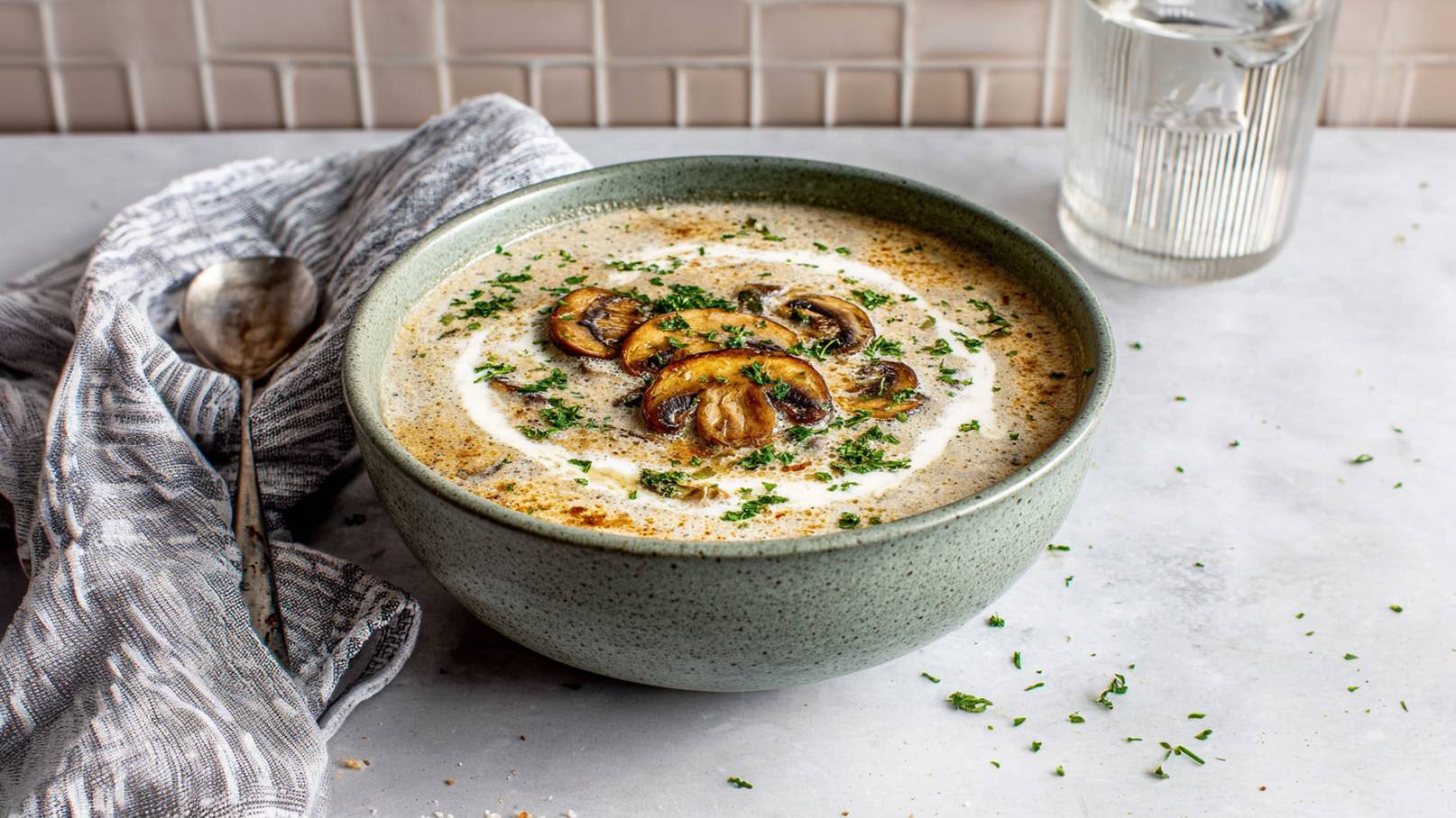 Eine grüne Schüssel mit cremiger Pilzsuppe, garniert mit Scheiben gebratener Pilze und einem Schuss Sahne. Fein gehackte Petersilie ist darüber gestreut. Im Hintergrund liegt ein Löffel und ein Glas Wasser steht daneben.