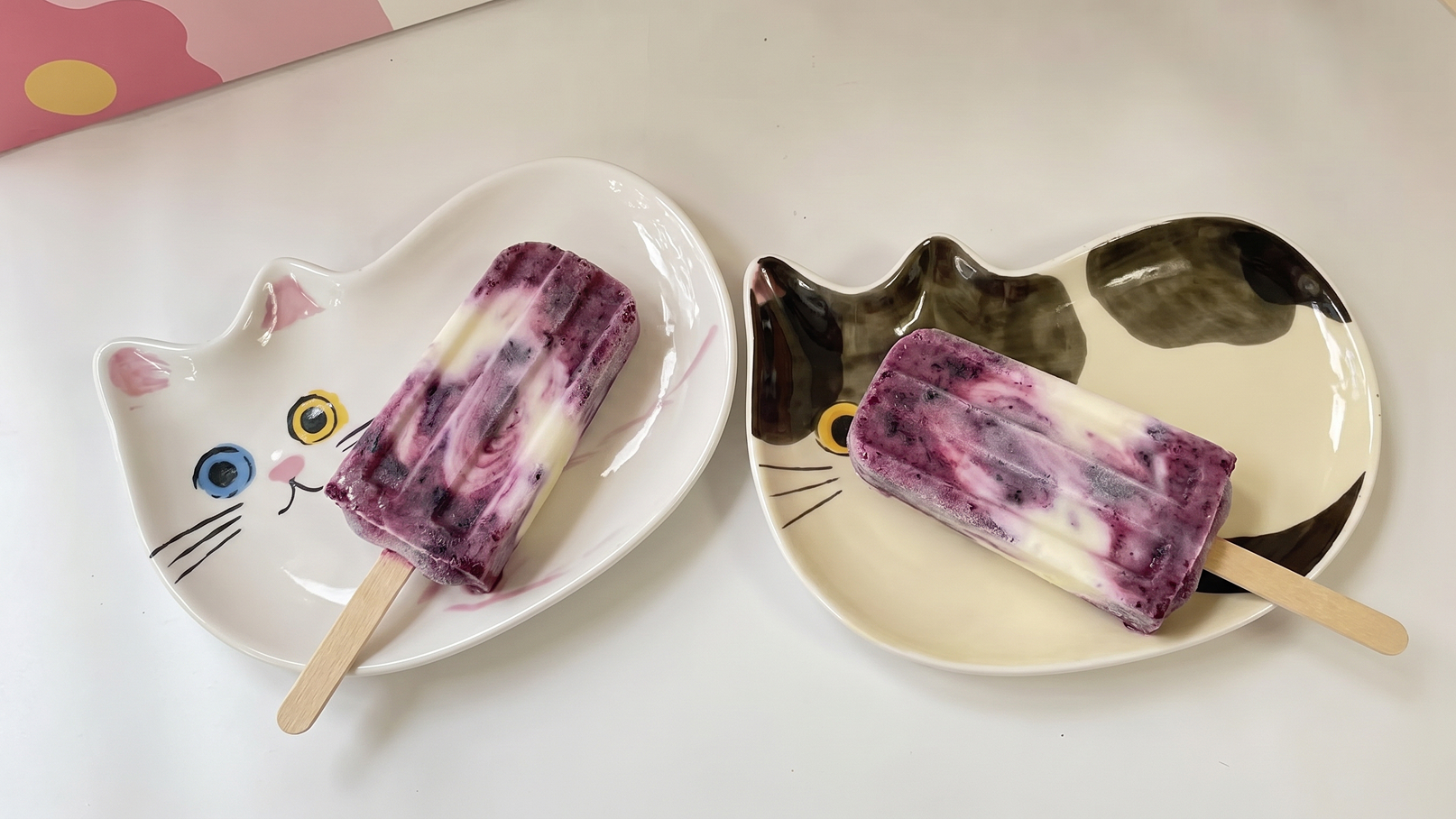 Zwei Eis am Stiel liegen auf Katzenförmigen Tellern. Das erste Eis ist lila-weiß marmoriert, das zweite ebenfalls mit einer ähnlichen Farbgestaltung. Die Teller sind in Form von Katzenköpfen mit Augen und Schnurrhaaren gestaltet.