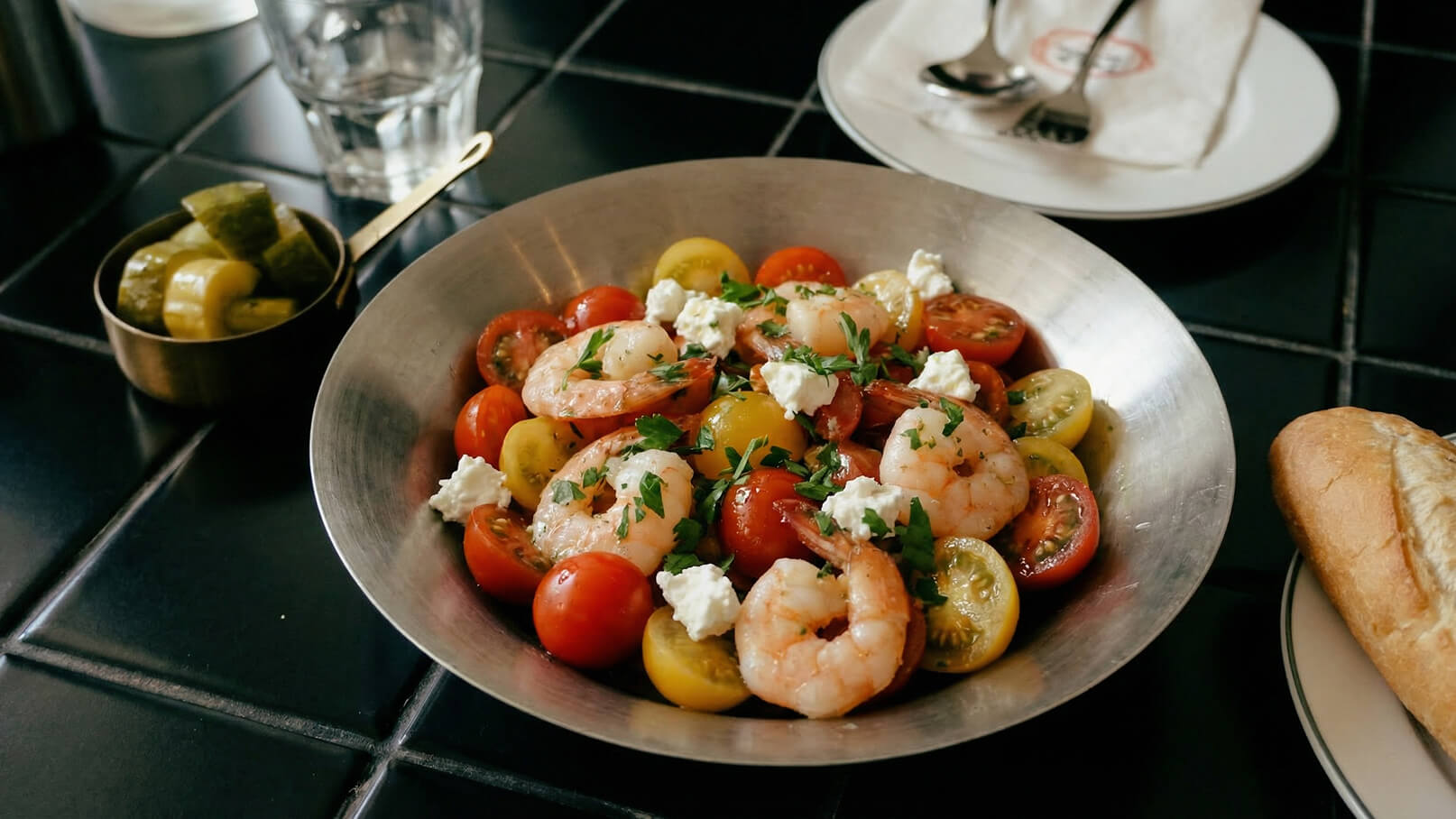 Ein silberner Teller mit einer bunten Mischung aus roten und gelben Tomaten, garniert mit großen, geschälten Garnelen und kleinen Stücken Frischkäse, die mit frischem Petersilie bestreut sind. Im Hintergrund sind eingelegte Gurken und ein Stück Brot zu sehen.