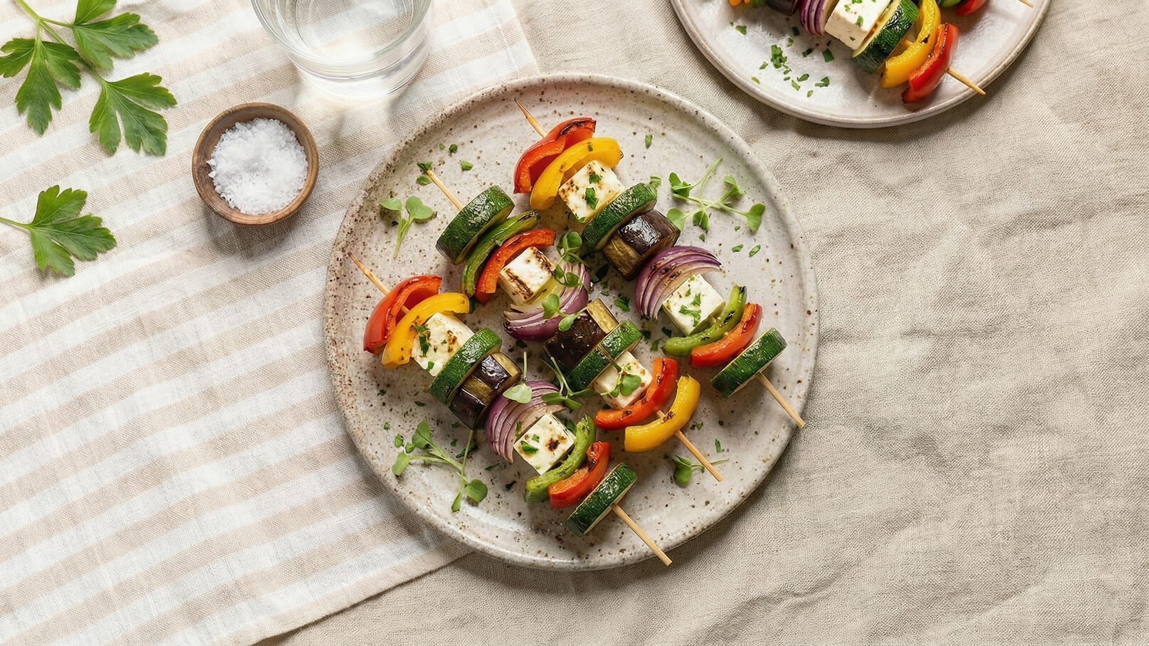 Ein Teller mit bunten Gemüsespießen, bestehend aus Zucchini, Paprika, Auberginen und Feta-Käse. Die Spieße sind mit frischen Kräutern garniert und auf einem hellen, strukturierten Tisch serviert. Ein Glas Wasser und eine Schale mit Salz sind ebenfalls sichtbar.