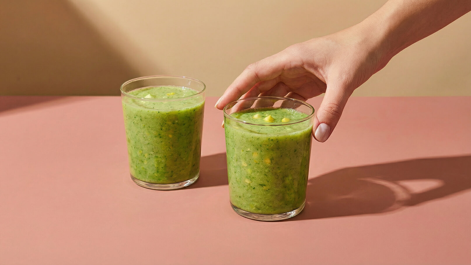Zwei Gläser mit grünem Smoothie stehen auf einem rosa Tisch. Eine Hand greift nach dem Glas rechts. Der Smoothie hat eine cremige Textur und ist mit kleinen Stückchen angereichert. Die Umgebung ist hell und einfach gestaltet.
