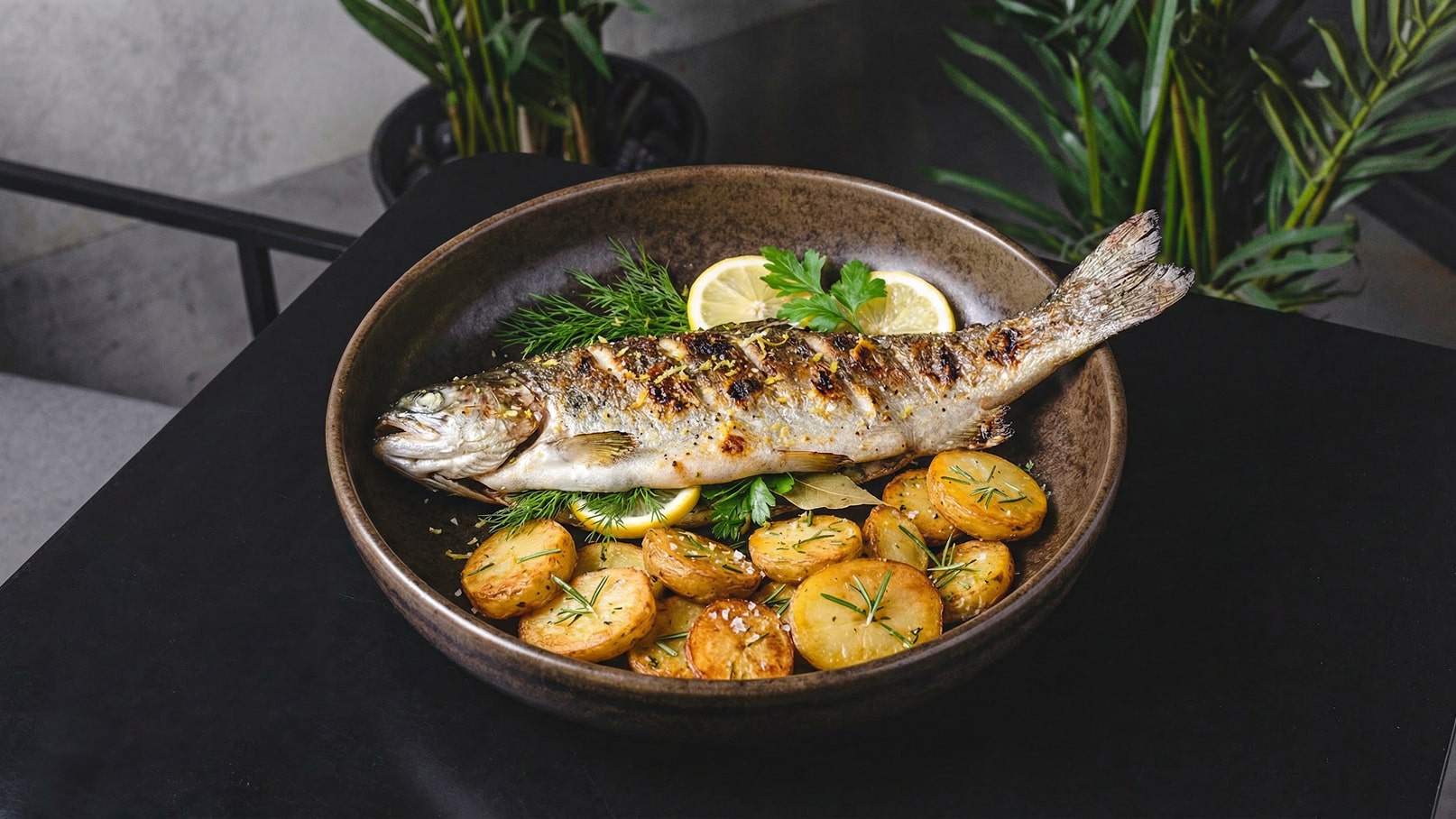 Auf dem Bild ist ein Teller mit einem gegrillten Fisch, umgeben von gebratenen Kartoffeln. Frische Kräuter, Zitronenscheiben und etwas Grünzeug sind ebenfalls zu sehen. Der Teller besteht aus dunklem Keramik und ist kunstvoll angerichtet.