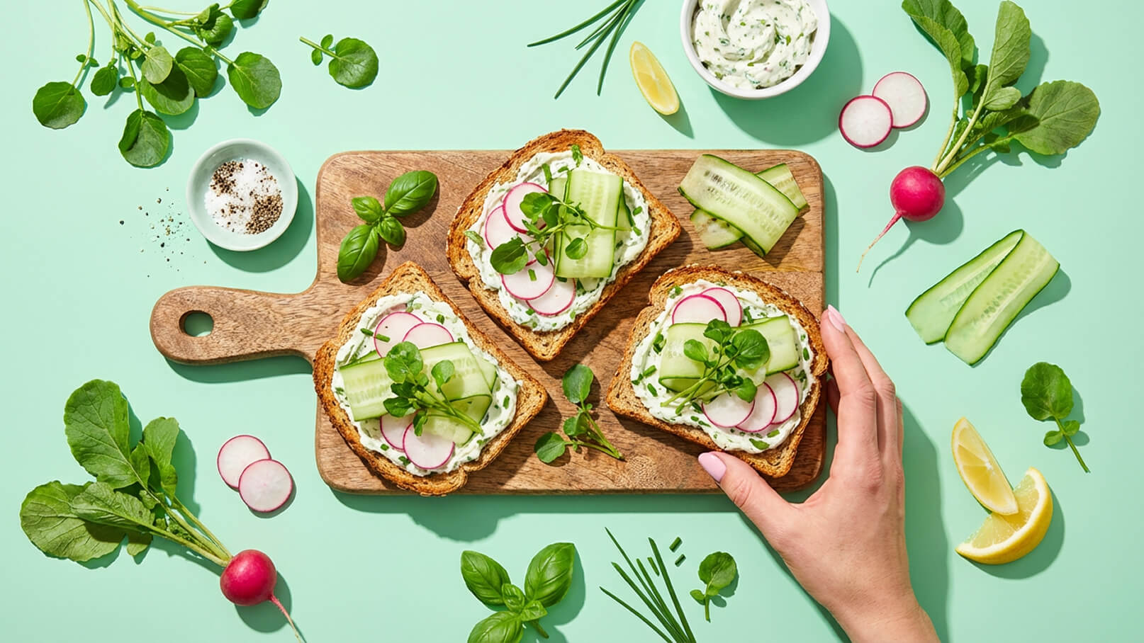 Auf einem Holzbrett liegen drei belegte Brote mit Frischkäse, Gurken und Radieschen. Daneben befinden sich frische Kräuter, Zitronenschnitze und Gewürze. Eine Hand greift nach einem der Brote. Der Hintergrund ist in einem sanften Grünton gehalten.