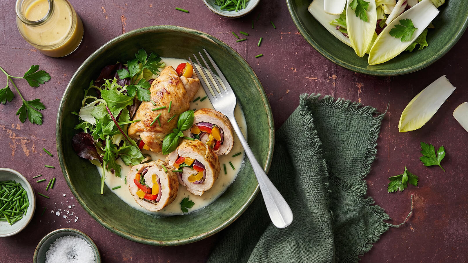 Ein Teller mit gefülltem Hähnchenrollbraten, serviert in einer cremigen Soße, garniert mit frischen Kräutern. Die Füllung besteht aus buntem Gemüse, sichtbar in aufgewickelter Form. Daneben befindet sich ein kleiner Salat aus blättrigem Gemüse und eine Glasflasche mit Dressing.