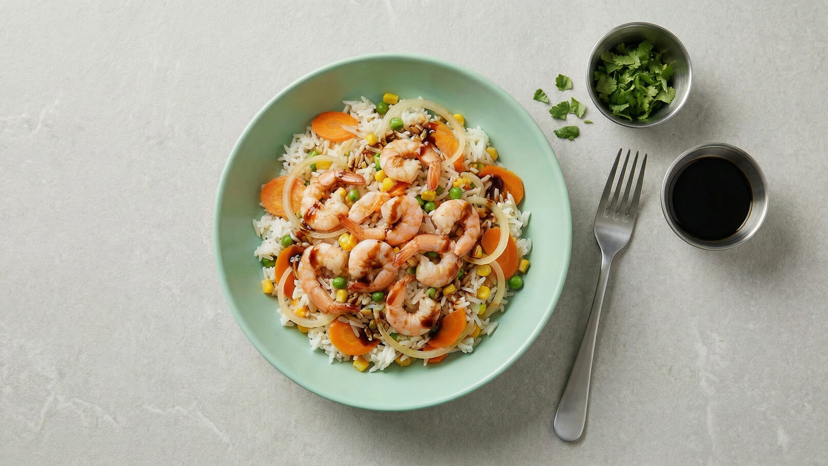 Ein Teller mit gebratenem Reis, überhäuft mit Garnelen, Karottenscheiben und Erbsen. Daneben befinden sich zwei kleine Schalen: eine mit frischen Korianderblättern und eine mit dunkler Sojasauce. Eine Gabel liegt ebenfalls neben dem Teller.