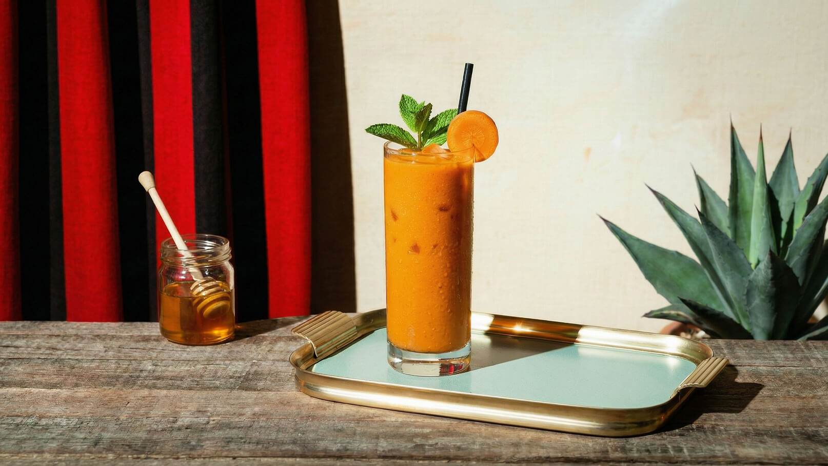 Ein hoher Glasbecher mit einem orangefarbenen Getränk, dekoriert mit einem Minzblatt und einer Orangenscheibe. Der Becher steht auf einem Tablett mit grünem Rand. Im Hintergrund ist ein Holztablett und eine Pflanze zu sehen.