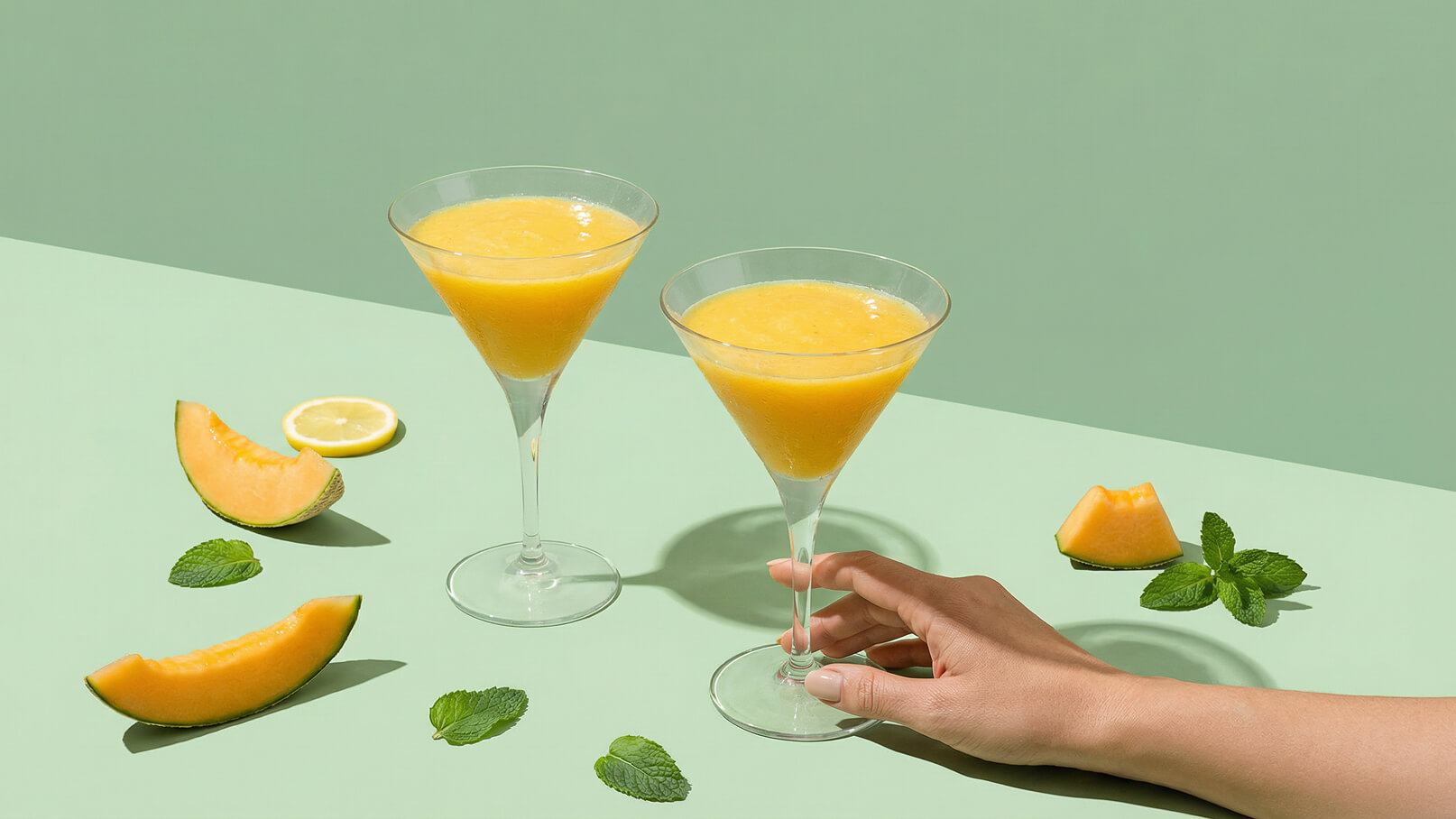 Zwei Gläser mit einem gelben Getränk stehen auf einer grünen Fläche. Eine Hand greift nach einem der Gläser. Um die Gläser liegen Melonenscheiben und frische Minzblätter.