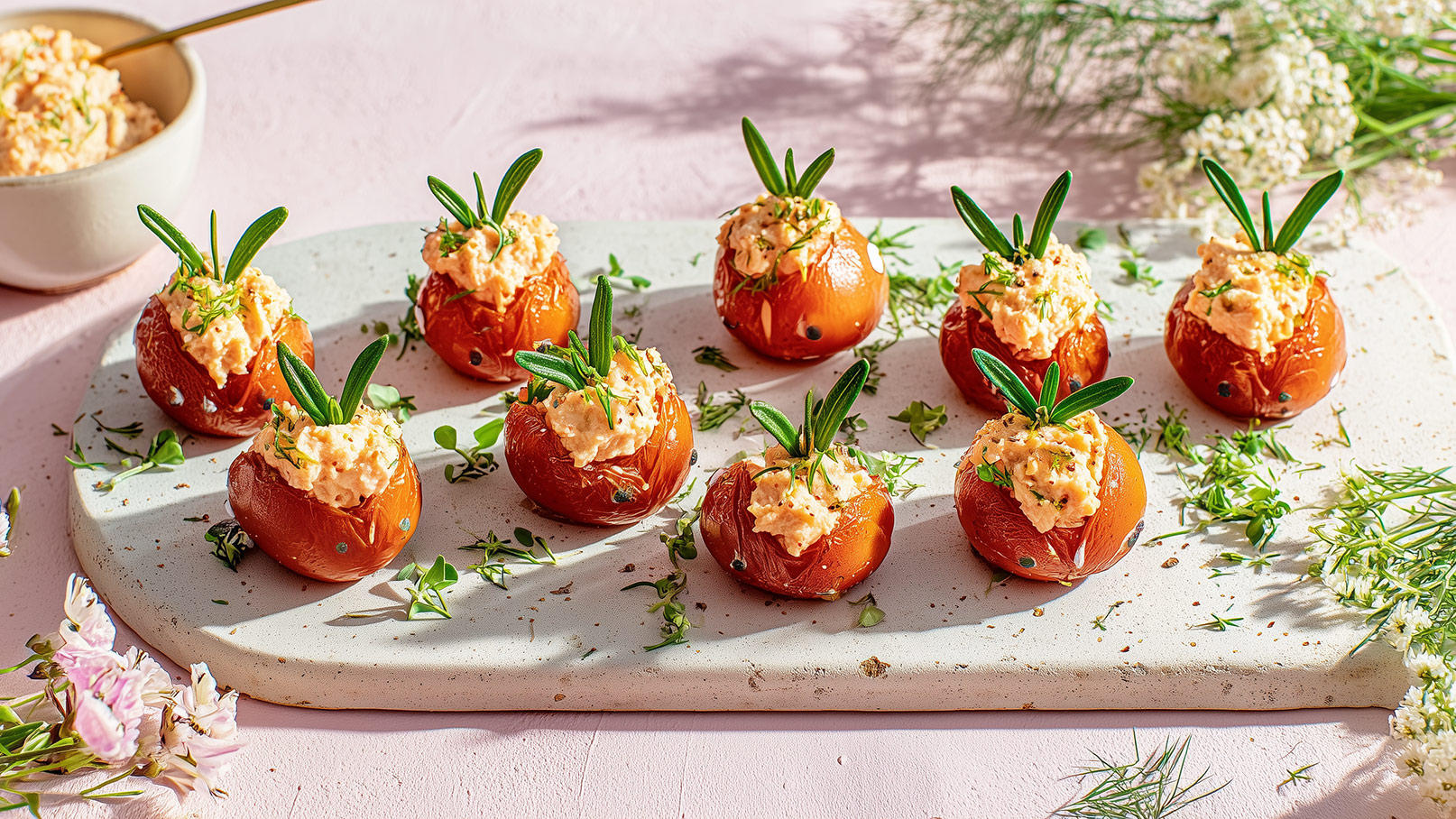 Ein Plattenteller zeigt gefüllte Tomaten, die mit einer cremigen Füllung und frischen Kräutern dekoriert sind. Der Hintergrund ist hell und es sind grüne Pflanzenteile um die Tomaten verteilt, die das Gericht ansprechend präsentieren.