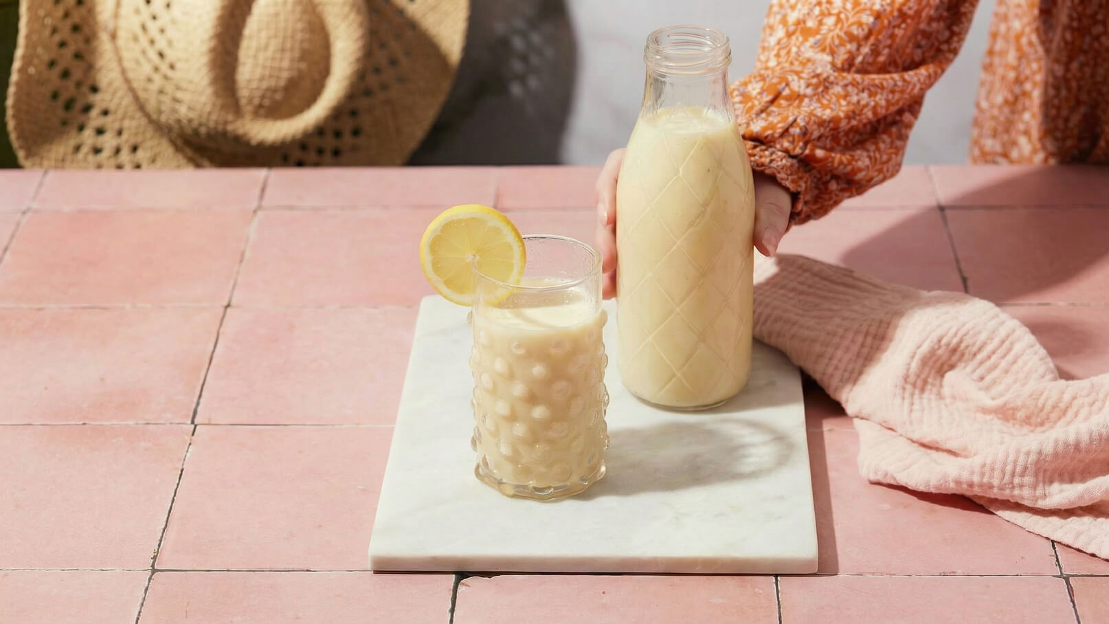 Ein Glas mit einem cremigen Getränk, garniert mit einer Zitronenscheibe, steht auf einem marmorierten Untersetzer. Daneben hält eine Hand eine Flasche mit der gleichen Flüssigkeit. Der Hintergrund zeigt eine rosa Fliesenoberfläche.