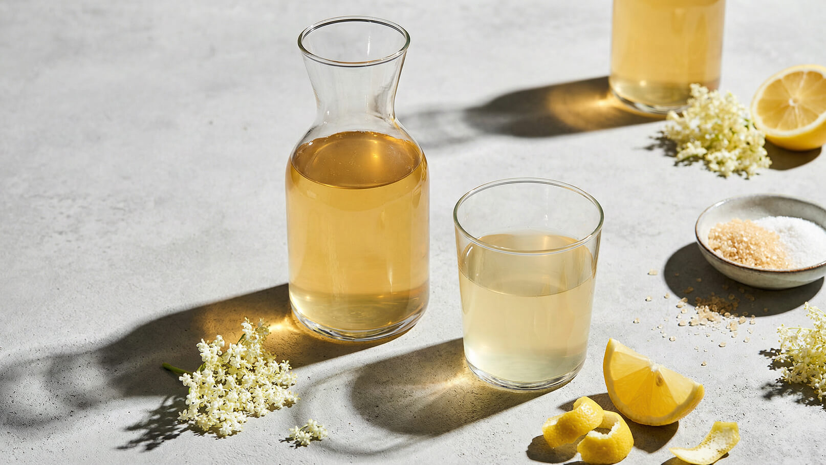 Ein Glaskrug mit einem hellbraunen Getränk steht neben einem Glas mit einer ähnlichen Flüssigkeit. Umgeben von Holunderblüten, Zitronenscheiben und Zucker, vermittelt die Anordnung eine erfrischende botanische Atmosphäre.