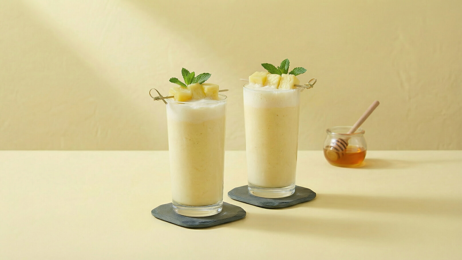 Zwei hohe Gläser mit einem cremigen, hellen Getränk, garniert mit Ananasstückchen und Minzblättern. Sie stehen auf schwarzen Untersetzern auf einem gelben Tisch. Im Hintergrund ist ein Glas Honig mit einem Holzlöffel zu sehen.