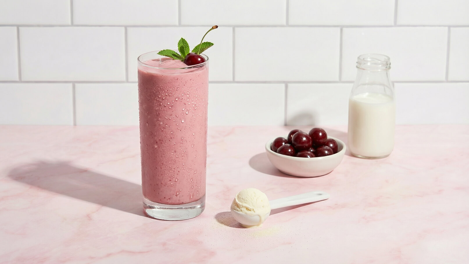 Ein hoher Glaskelch mit einem rosa Smoothie, garniert mit einem Blatt Minze und einer Kirsche, steht auf einem hellrosa Tisch. Neben dem Glas befindet sich ein Becher mit Kirschen und eine Flasche Milch. Ein Löffel mit einer Portion Eiscreme liegt nahe dem Glas.