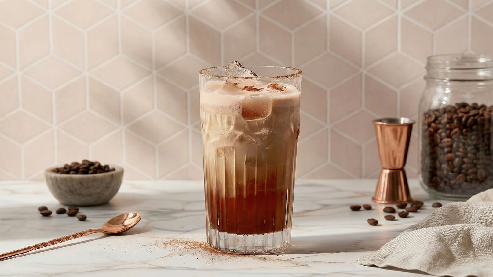 Ein Glas mit eisgekühltem Kaffee, der geschichtet ist, steht auf einem marmorierten Tisch. Die obere Schicht ist cremig und hell, die untere dunkel. Darum sind Kaffeebohnen und ein Messbecher angeordnet, die das Kaffeethema unterstreichen. Die Hintergrundfarbe ist sanft und geometrisch.