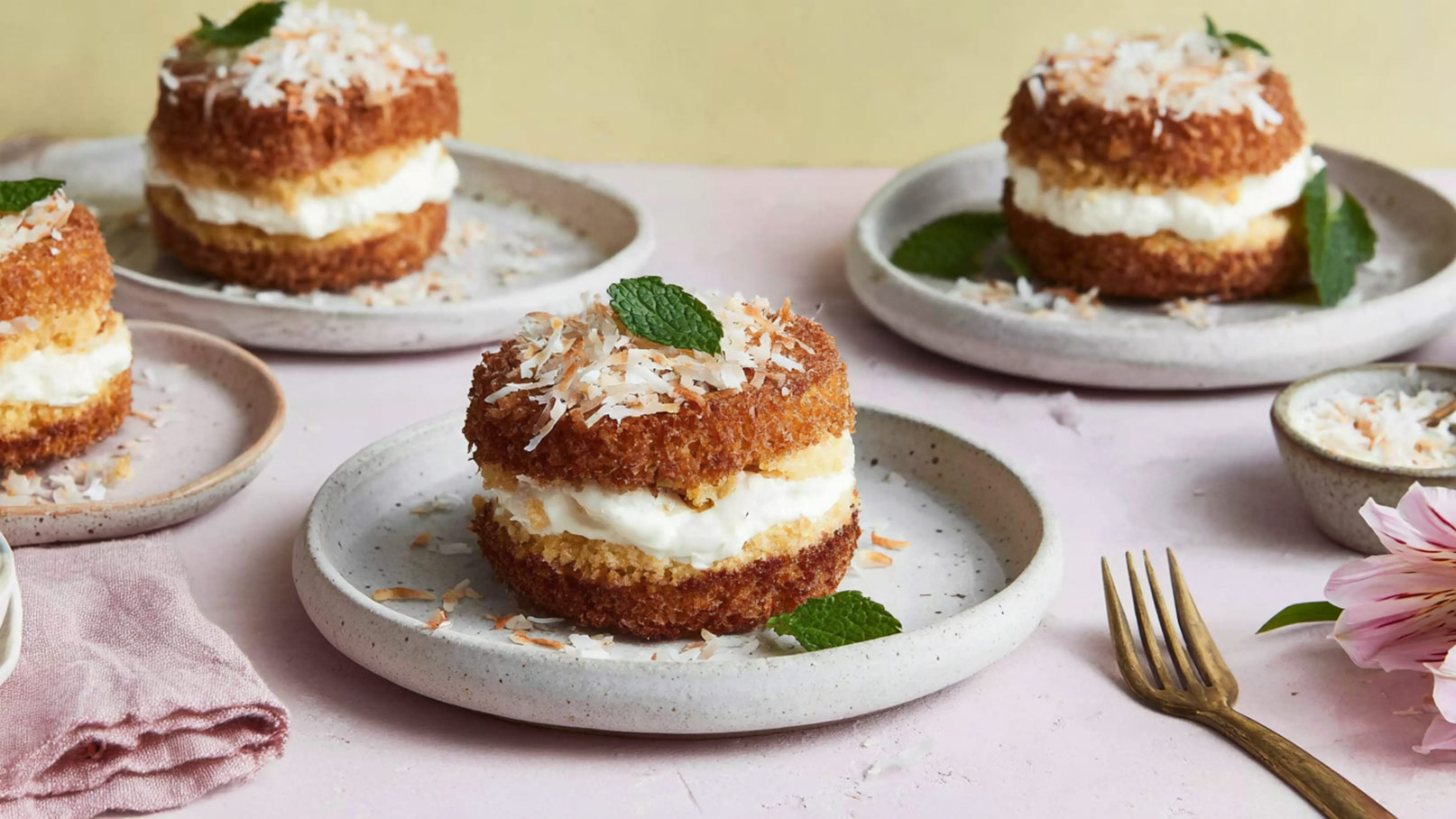 Verschiedene Stücken von Kokoskuchen auf hellen Tellern, angerichtet mit Sahnefüllung und Kokosraspeln. Einige Platten haben frische Minzblätter und Kokosraspeln daneben. Der Untergrund ist rosa und passend zur Präsentation des Desserts.