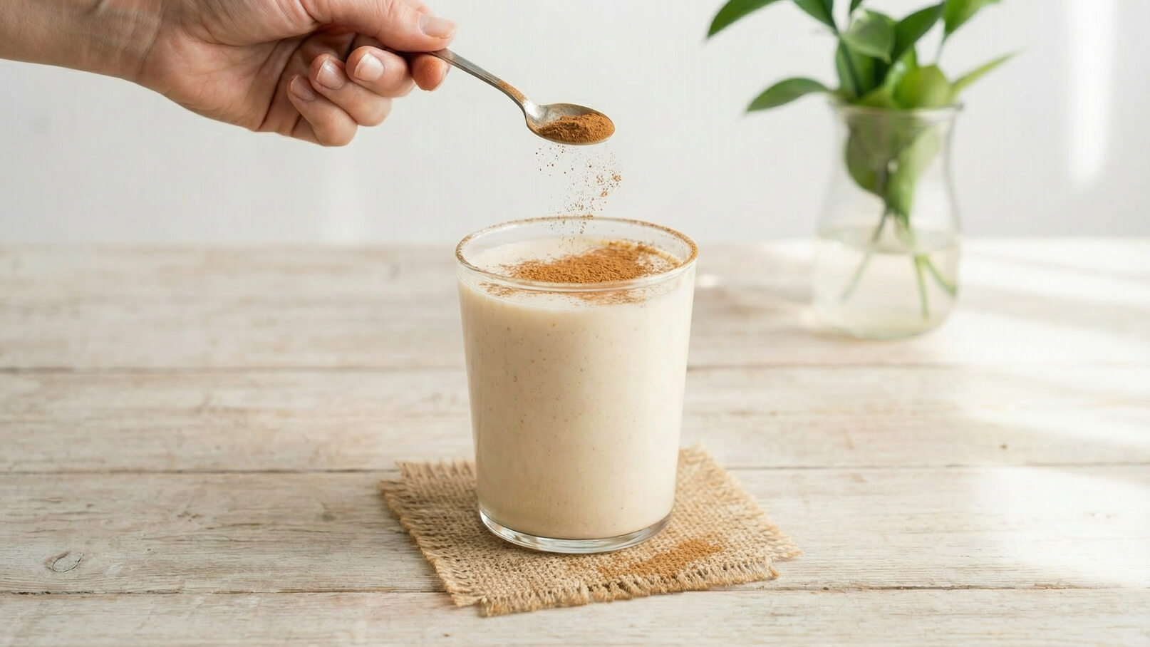 Eine Hand hält einen Löffel mit Gewürz, während sie es über ein Glas mit cremigem Getränk streut. Das Glas steht auf einem natürlichen Untersetzer. Im Hintergrund ist eine Vase mit Pflanzen sichtbar.