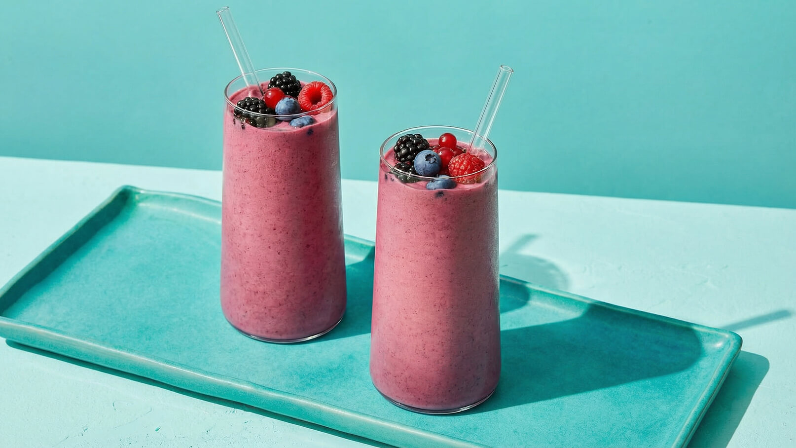 Zwei hohe Gläser mit einem pinken Smoothie stehen auf einem hellblauen Tablett. Die Smoothies sind mit verschiedenen Beeren, darunter Himbeeren, Blaubeeren und Brombeeren, dekoriert und haben jeweils einen Strohhalm. Der Hintergrund ist ein sanftes Blau.